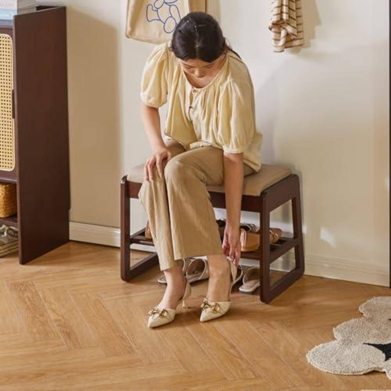Frau sitzt auf einer Bank und verstellt ihren beigefarbenen Schuh in einem minimalistischen Eingangsbereich mit Schuhen, die darunter gelagert sind.