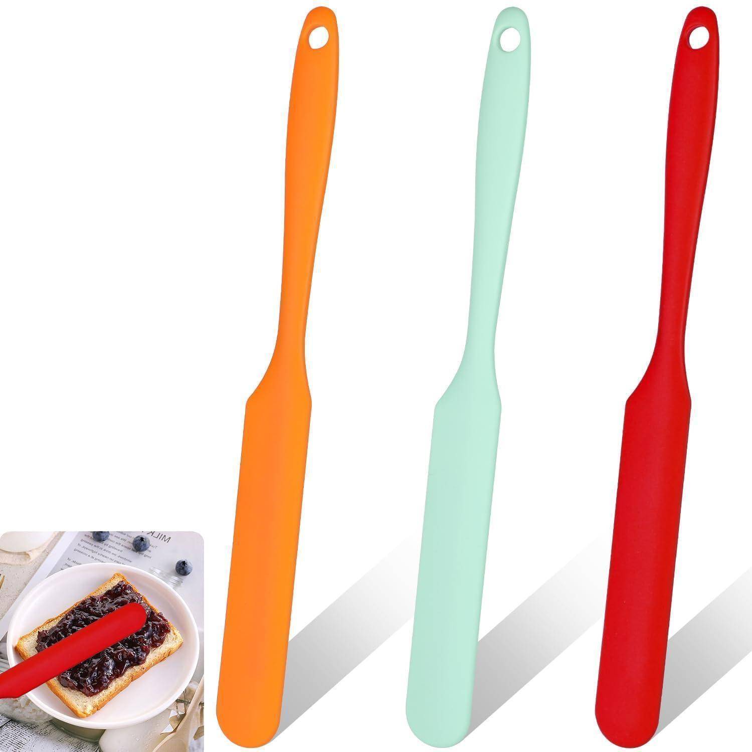 Drei farbenfrohe Silikon-Speichelmesser mit Aufhängelöchern, gezeigt in Orange, Mintgrün und Rot; die Vergrößerung zeigt eines, das Marmelade auf Toast verteilt.