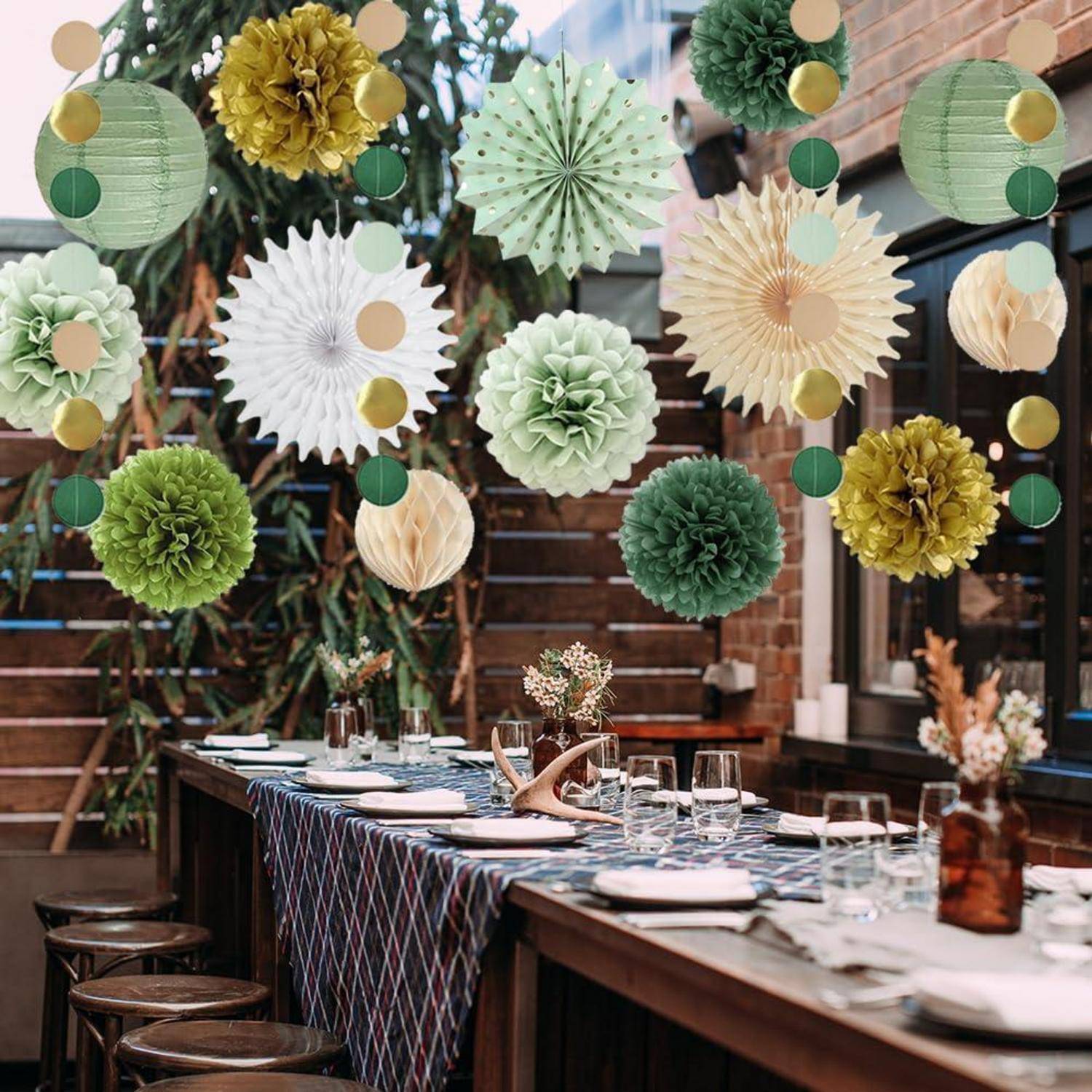 Festlich gedeckter Esstisch für eine Party, mit hängenden grünen, gold- und weißfarbenen Papierrosetten über den Gedeck in einem Patio oder Gartenbereich.