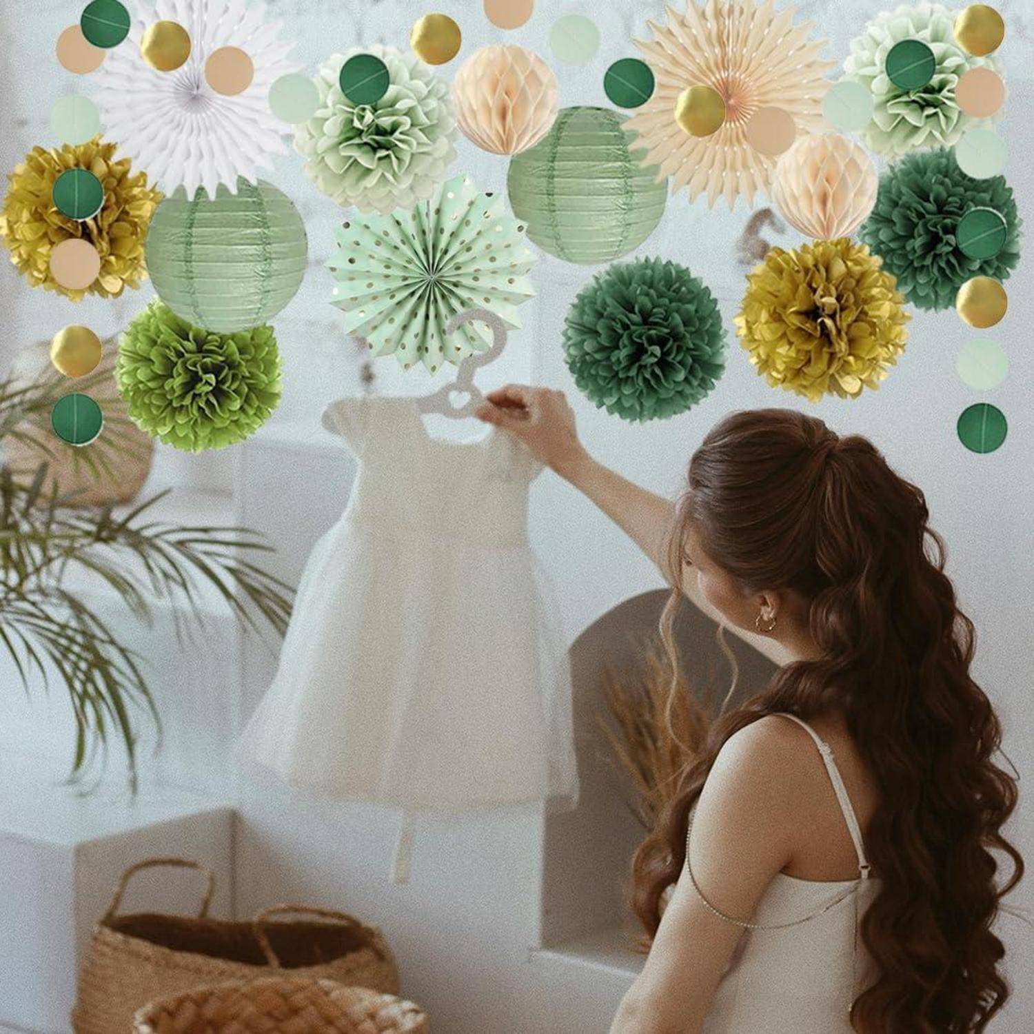 Frau in einem weißen Kleid stellt ein kindergrößes weißes Kleid auf einer Schneiderpuppe ein, umgeben von grünen, goldenen und cremefarbenen Papierdekoration.