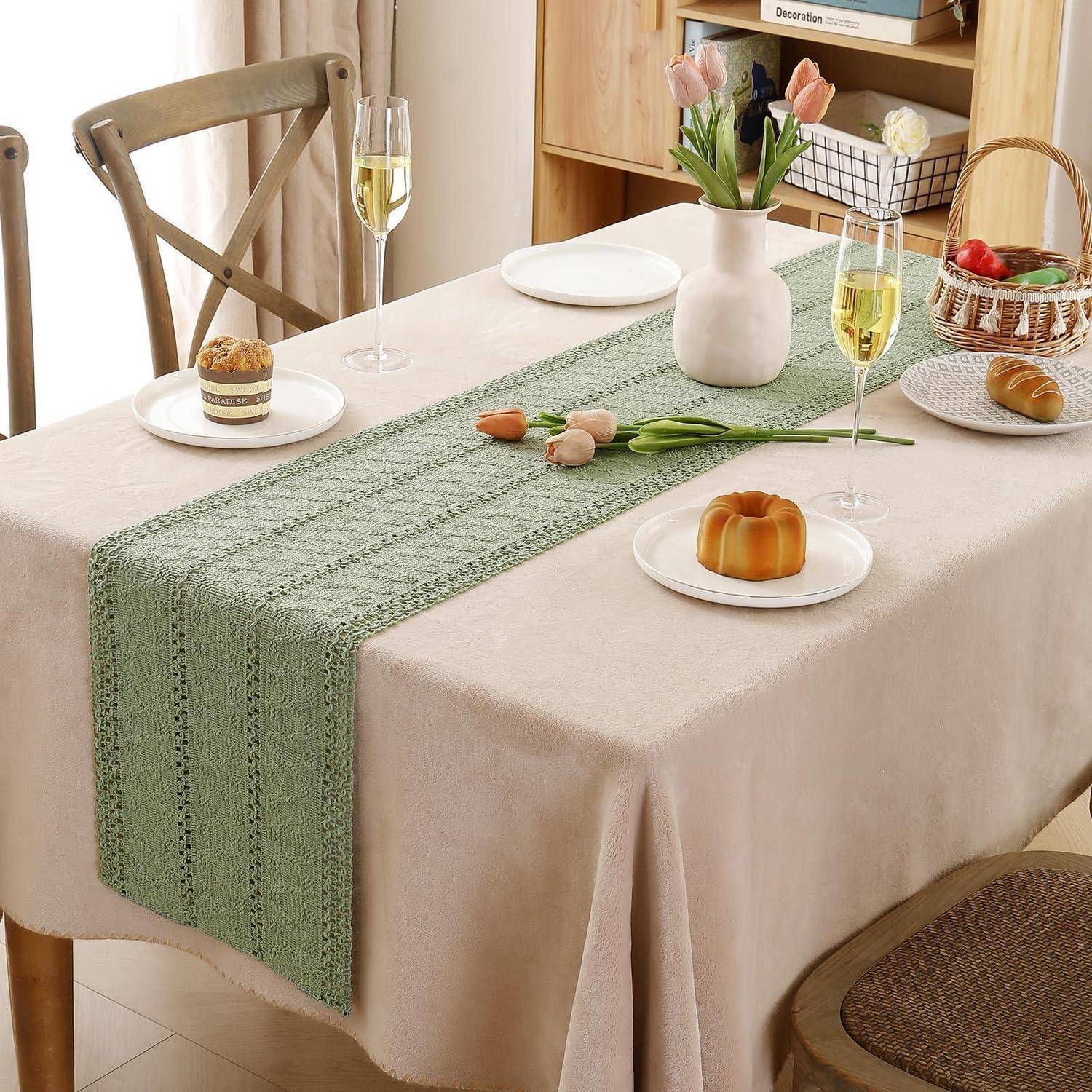 Esstisch mit beigefarbiger Tischdecke und grünem gewebtem Tischläufer, eingedeckt mit Gebäck, Champagnergläsern, Tulpen in einer Vase und Tellern in einem hellen Zimmer.