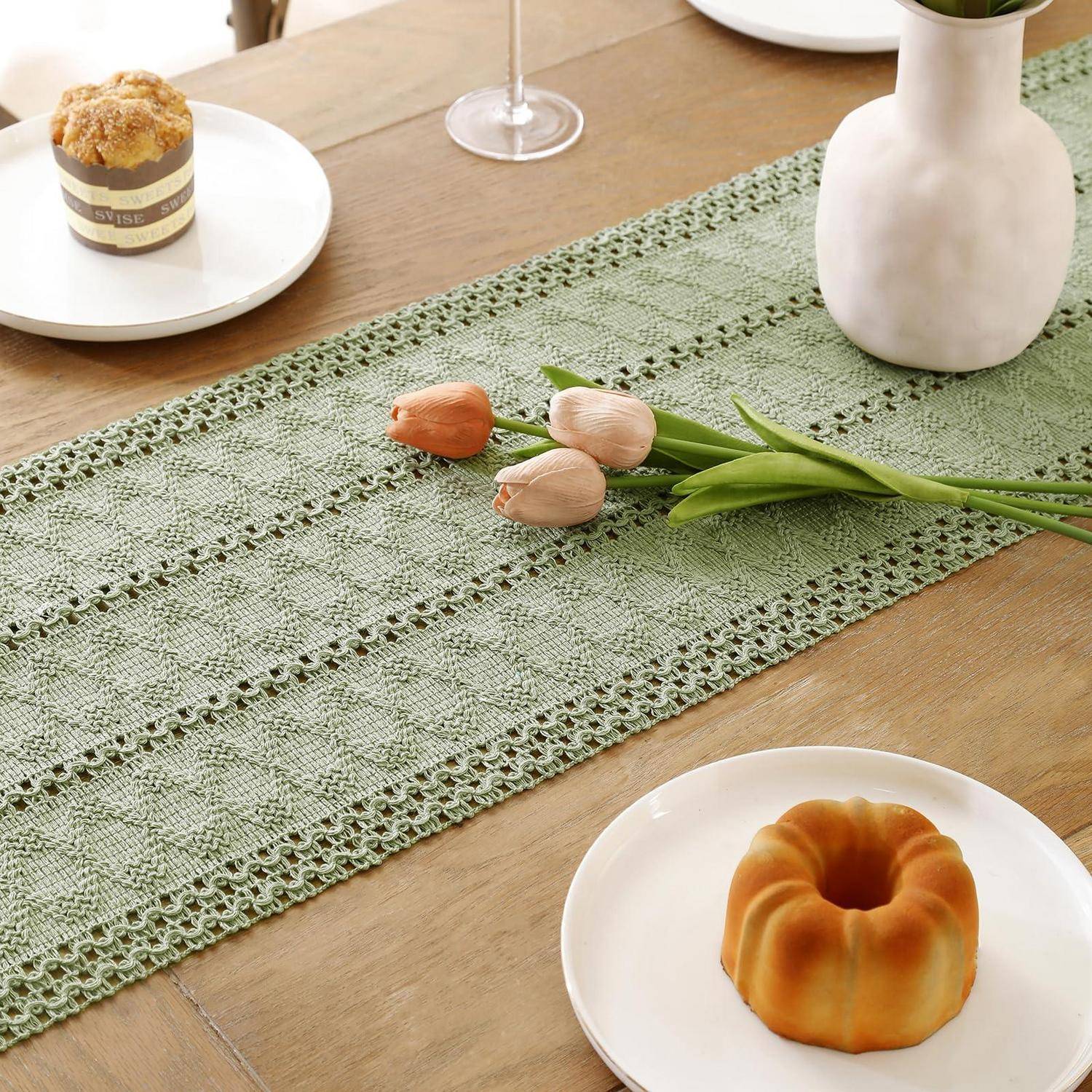 Tischgedeck mit einem hellgrünen Spitzenlaufer, Tulpen in der Mitte, einer weißen Vase und kleinen Kuchen auf weißen Tellern.