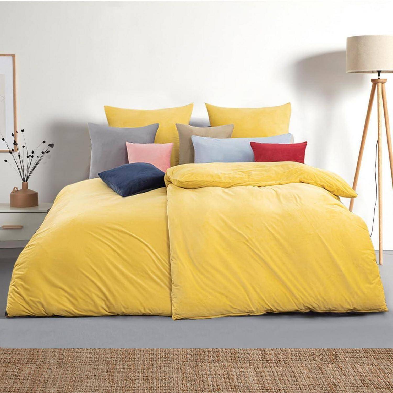 Ein helles gelbes Bett mit mehreren bunten Kissen und einer Stehleuchte in einem minimalistischen Schlafzimmer.