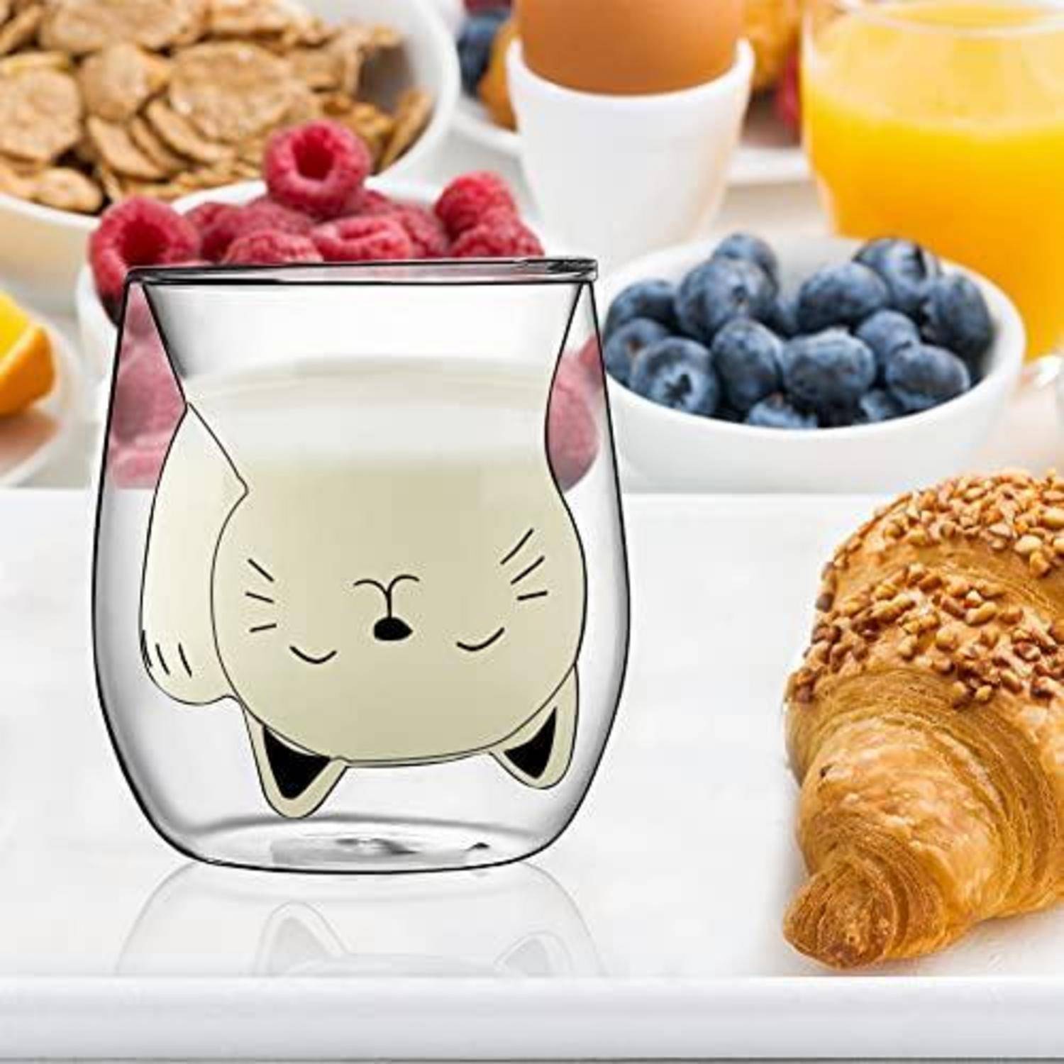 Doppelwandige Glastasse mit schlafendem Katzengesicht-Design auf einem Frühstückstisch neben Beeren, Müsli, Saft und einem Croissant.