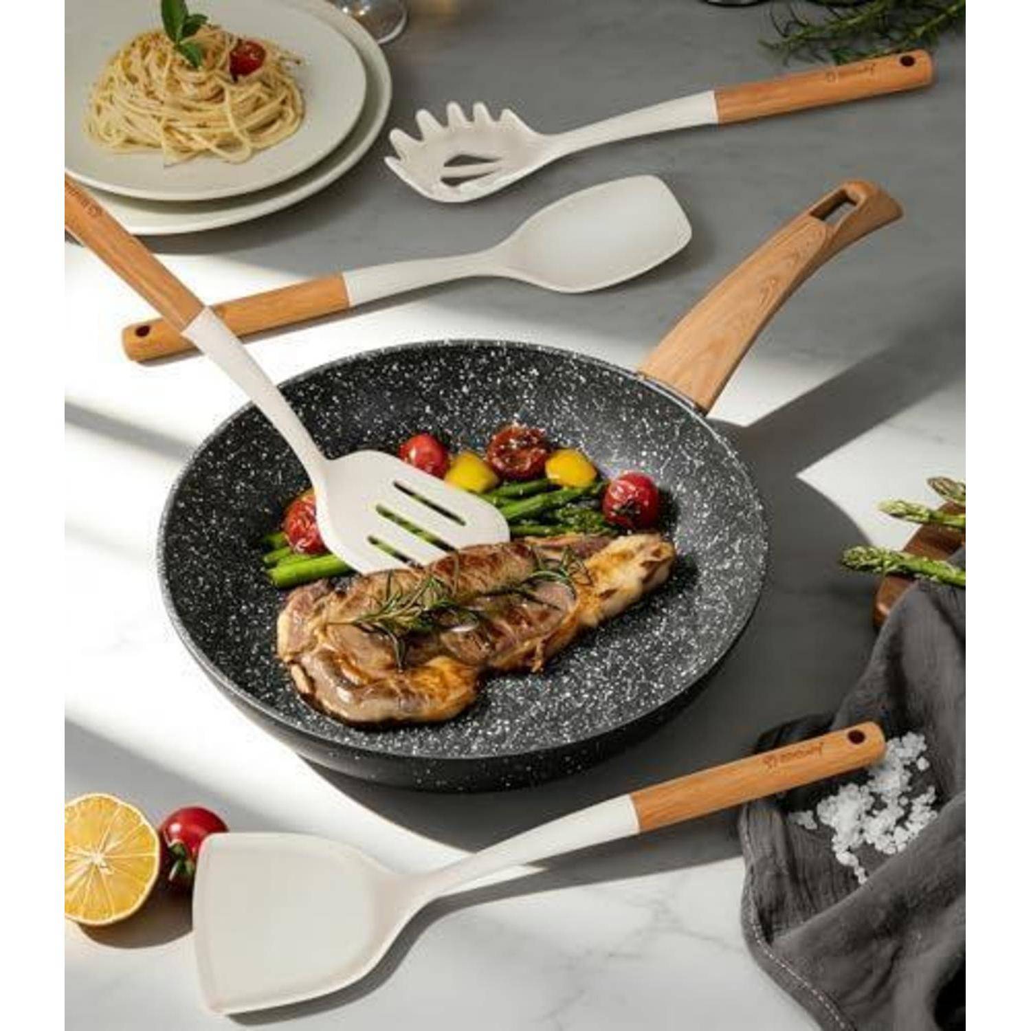 Gegrilltes Steak in einer gesprenkelten Bratpfanne mit Spargel und Gemüse, serviert mit Nudeln auf einem Teller und Küchenutensilien rundherum.