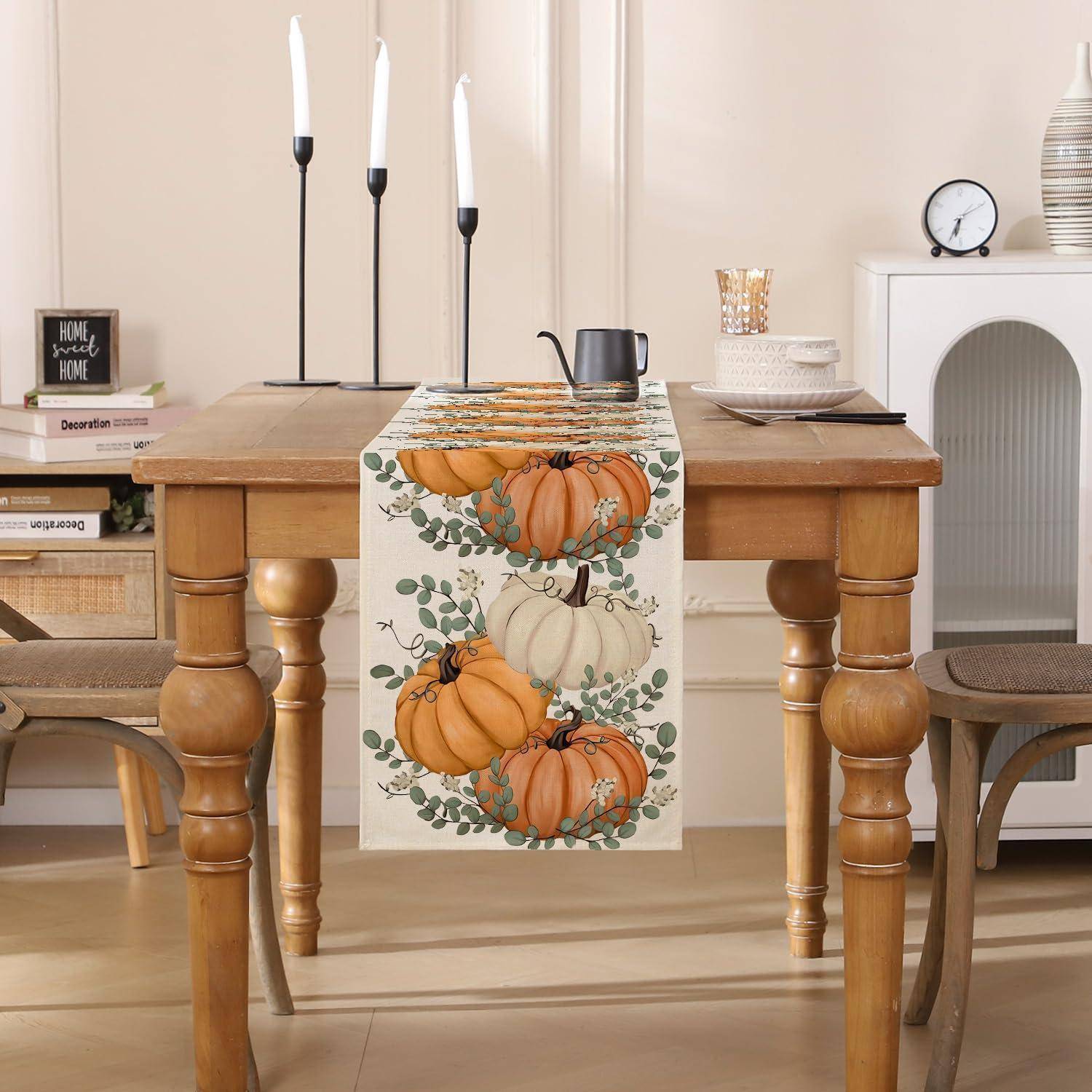 Holz-Esstischset für den Herbst, mit einem Tischläufer mit Kürbismuster, Kerzenleuchtern, Tischbesteck und Wohndekoration in einem gemütlichen beigen Zimmer.