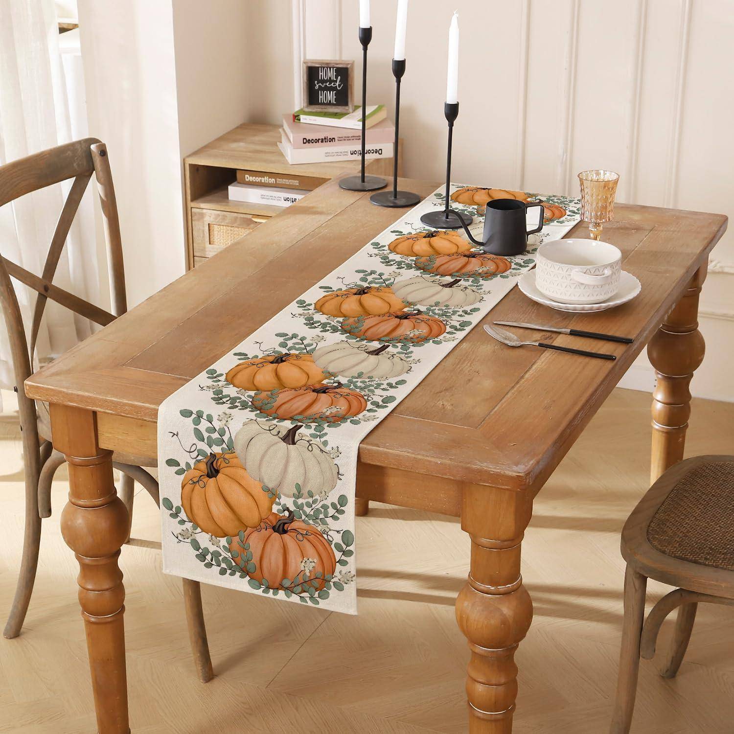 Holztisch-Esszimmerset für eine Herbstmahlzeit, mit Kürbis-Tischläufer, Kerzen, Tellern, Tassen und Stühlen in einem hellen Raum.