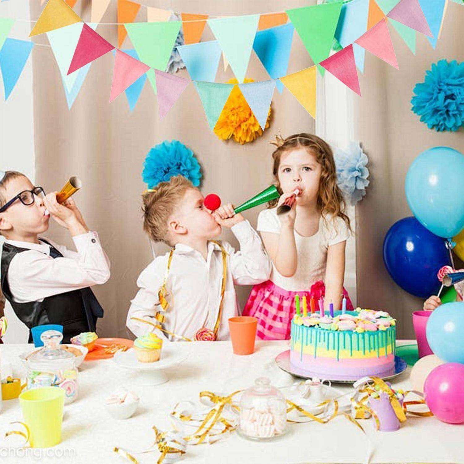 Drei Kinder in Partykleidung blasen in Partytröten an einem dekorierten Geburtstagstisch mit Kuchen, Ballons und Girlanden.