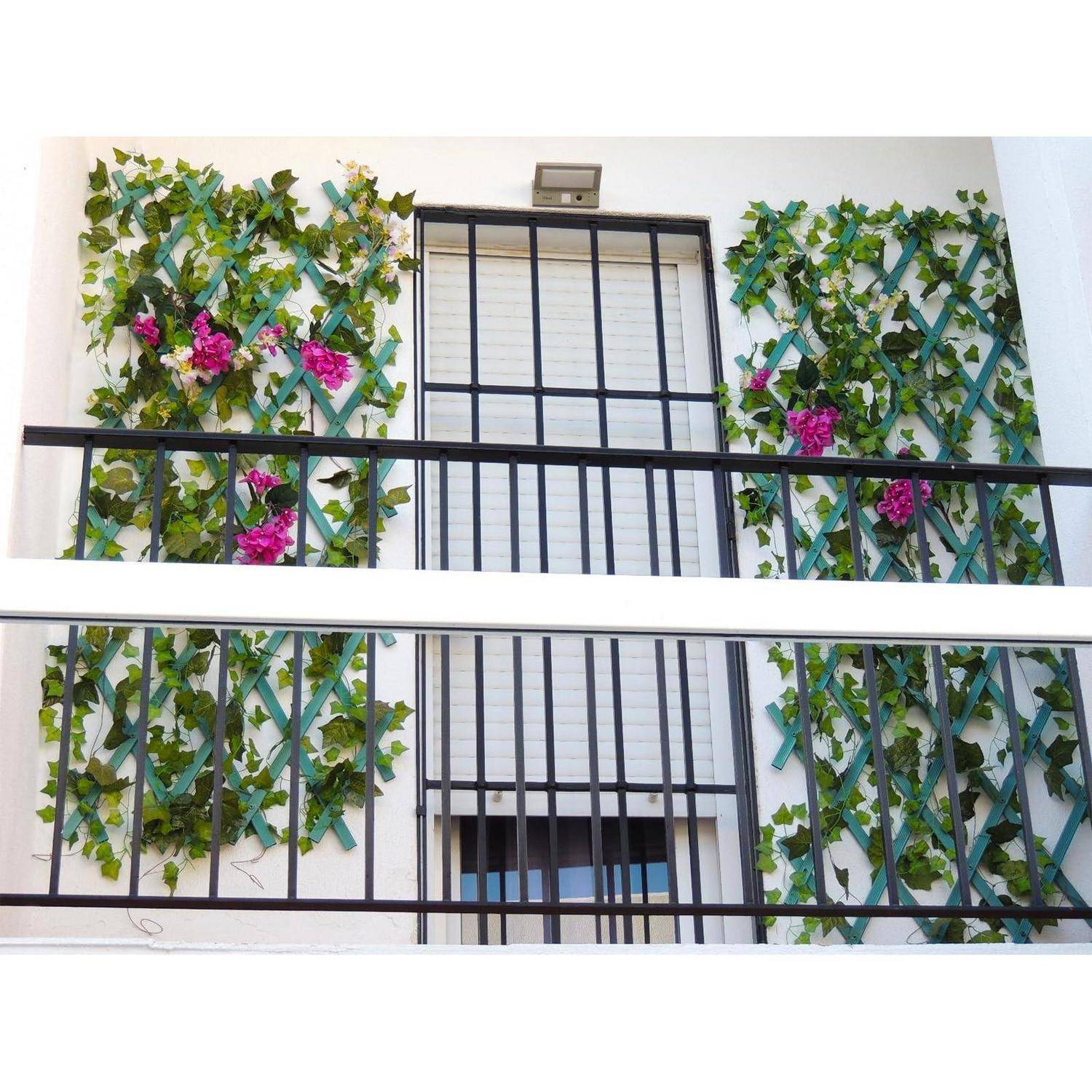Balkon einer Wohnung mit schwarzem Geländer, verbarrikadiertem Fenster und dekorativen grünen Ranken mit rosa Blüten auf beiden Seiten.