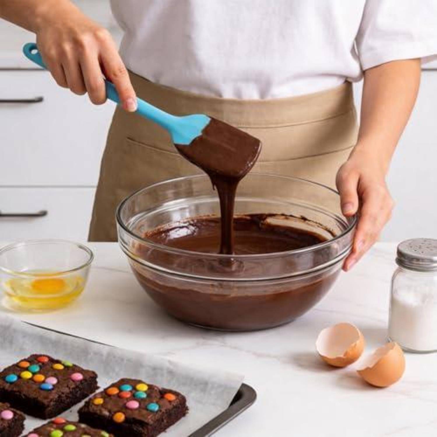 Eine Person rührt geschmolzene Schokolade in einer Schüssel um, während sie Brownies backt; Eier, Zucker und gebackene Brownies-Quadrate liegen auf der Arbeitsplatte.