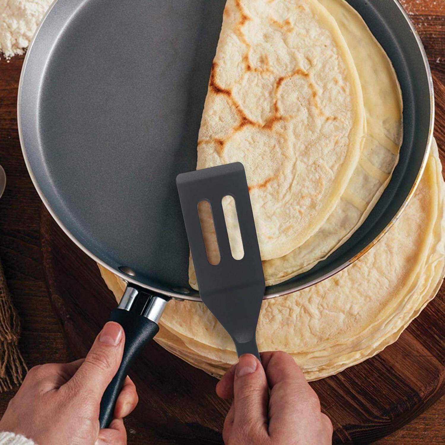 Hände verwenden einen Spatel, um einen Crêpe aus einer Bratpfanne auf einer Holzoberfläche zu heben.