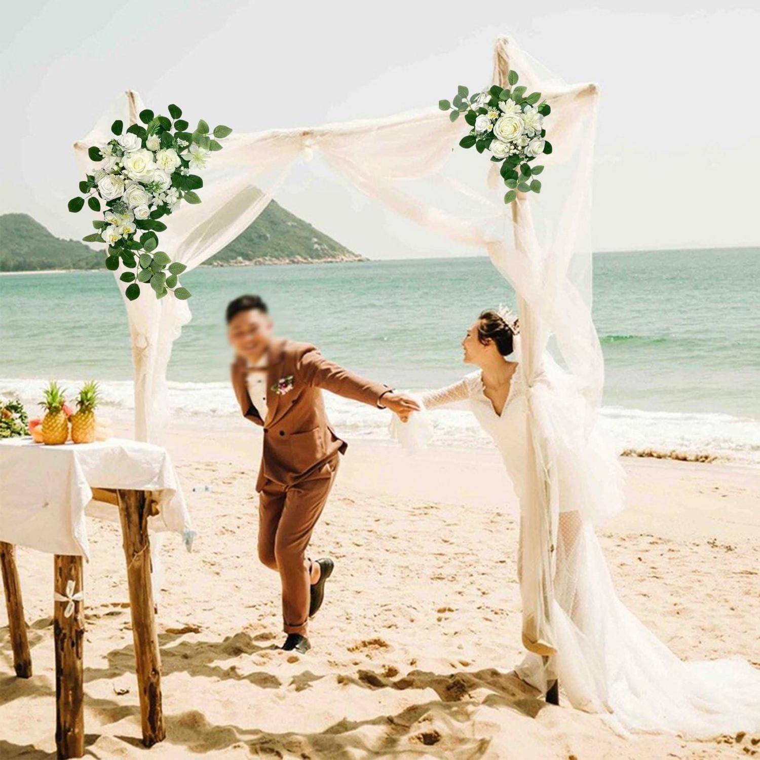 Ein Paar, das sich unter einem geschmückten Traubogen an der Hochzeit am Strand an den Händen hält, wobei die Braut in Weiß und der Bräutigam in einem braunen Anzug am Meer stehen.