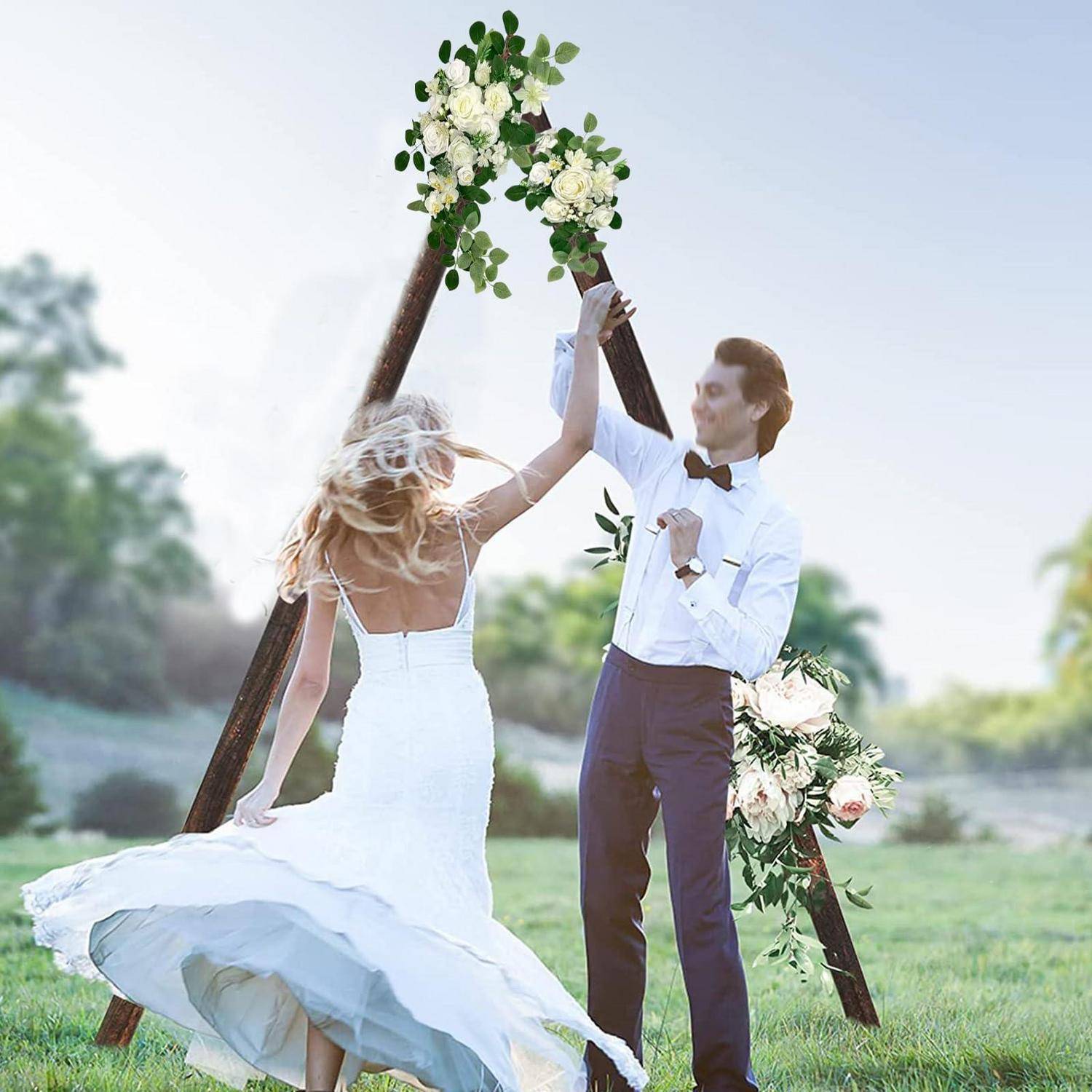 Braut in einem weißen Hochzeitskleid und Bräutigam in einem Smoking halten sich unter einem Blumenbogen im Freien am Wasser an den Händen.