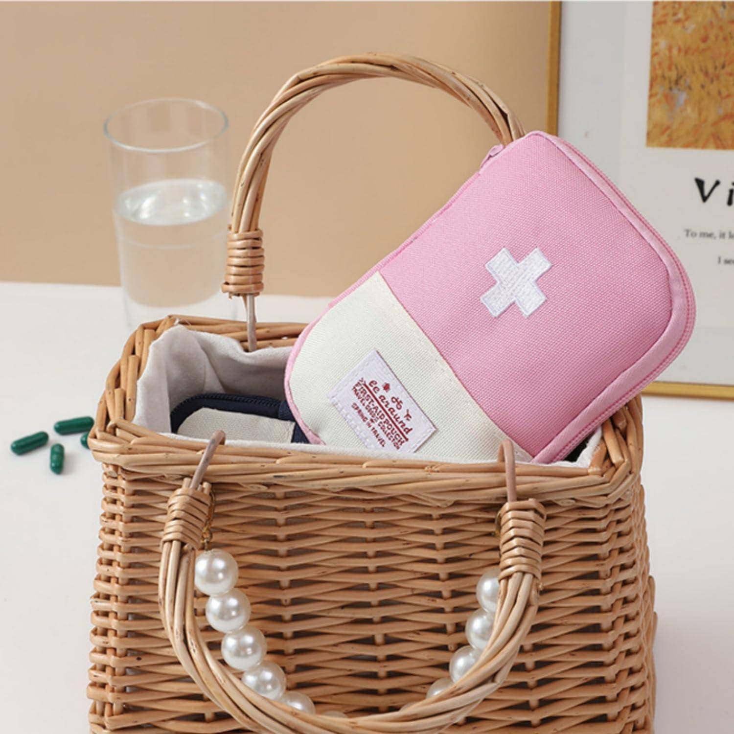 Rosa Erste-Hilfe-Kasten mit weißem Kreuz in einem Weidenkorb, mit Tabletten und einem Glas Wasser in der Nähe, was auf einen häuslichen Sanitätskasten hindeutet.