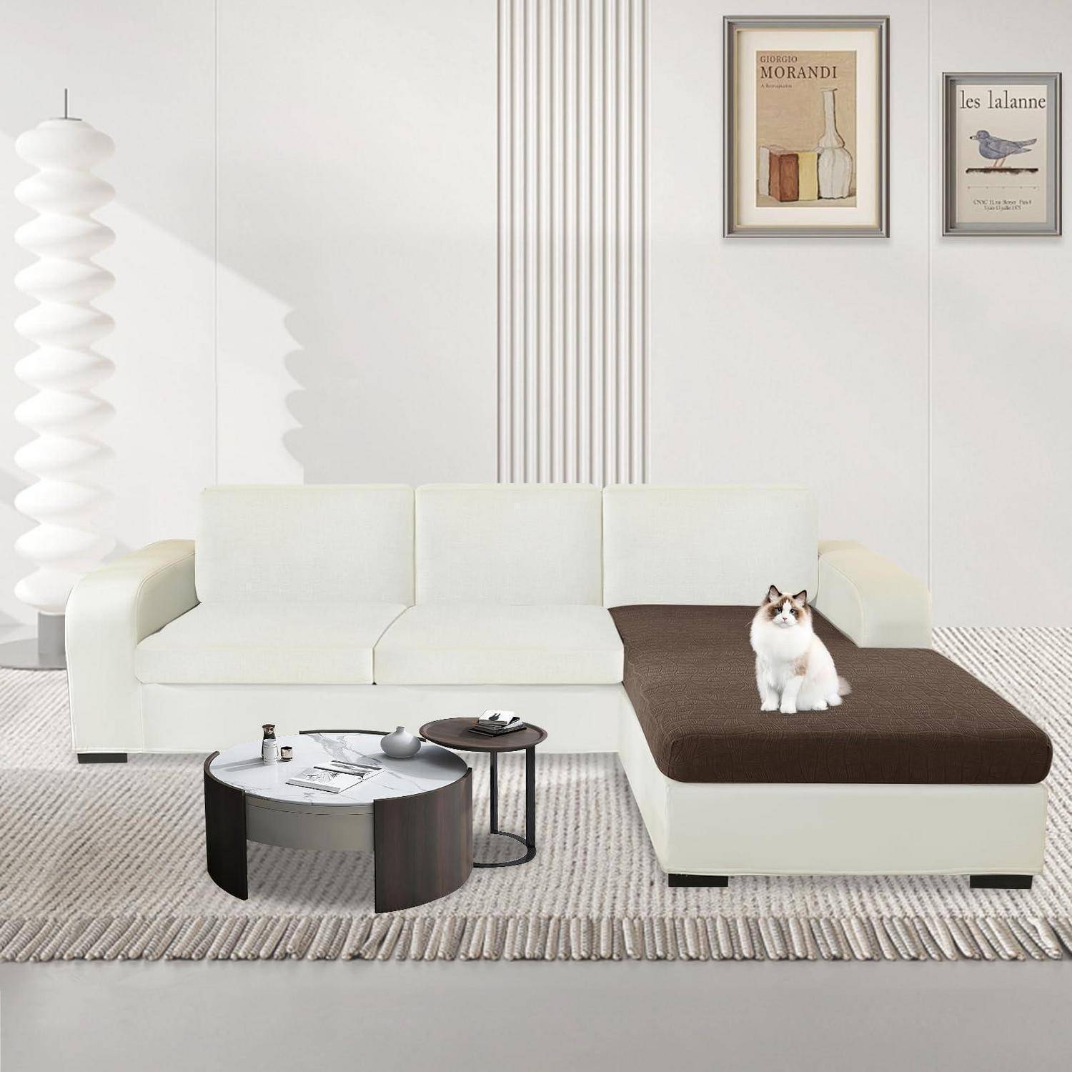 Eine Katze sitzt auf einem weißen Ecksofa in einem minimalistischen Wohnzimmer mit gerahmten Wandbildern, einer Stehlampe und einem runden Couchtisch auf einem Teppich.