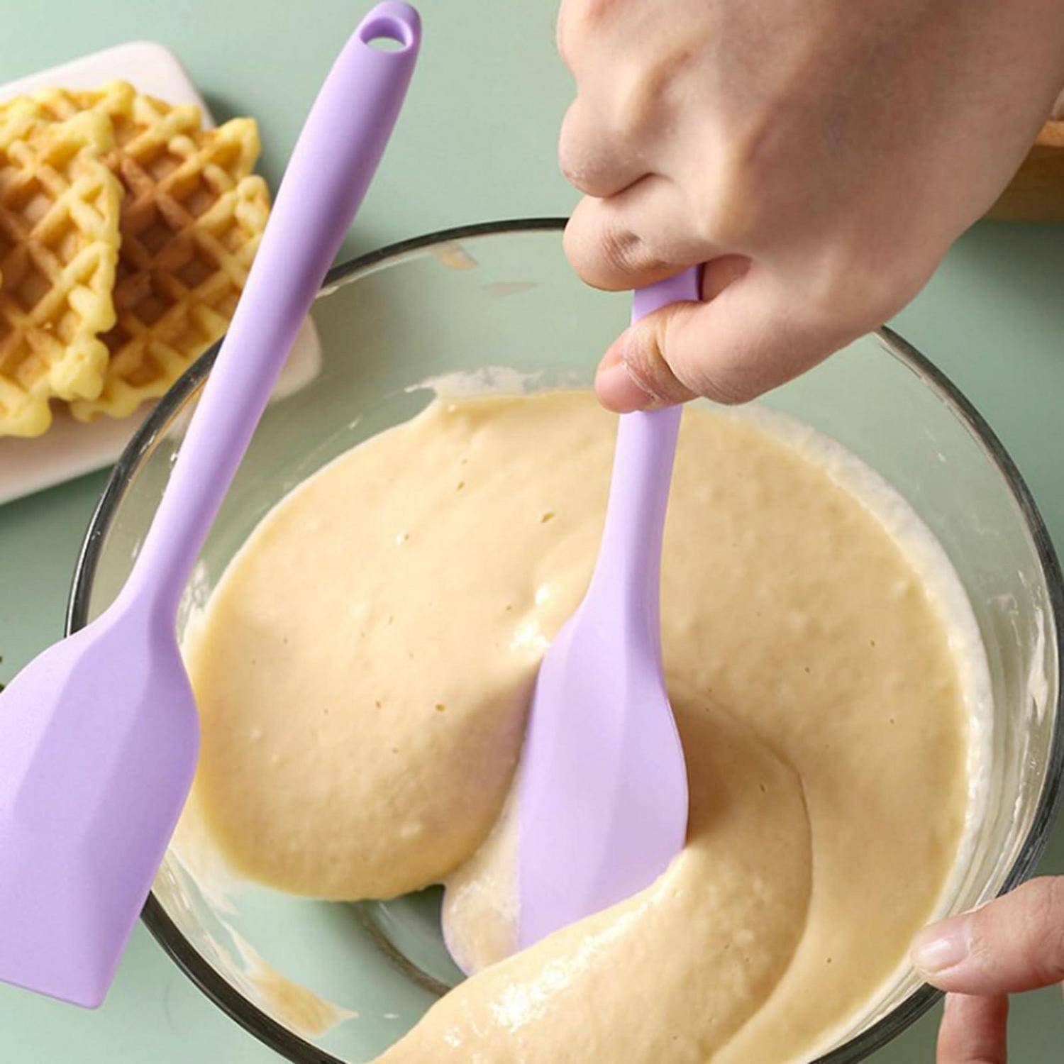 Mit der Hand dicken Teig in einer Glassschüssel mit einem violetten Spatel umrühren, neben einem Teller mit Waffeln.