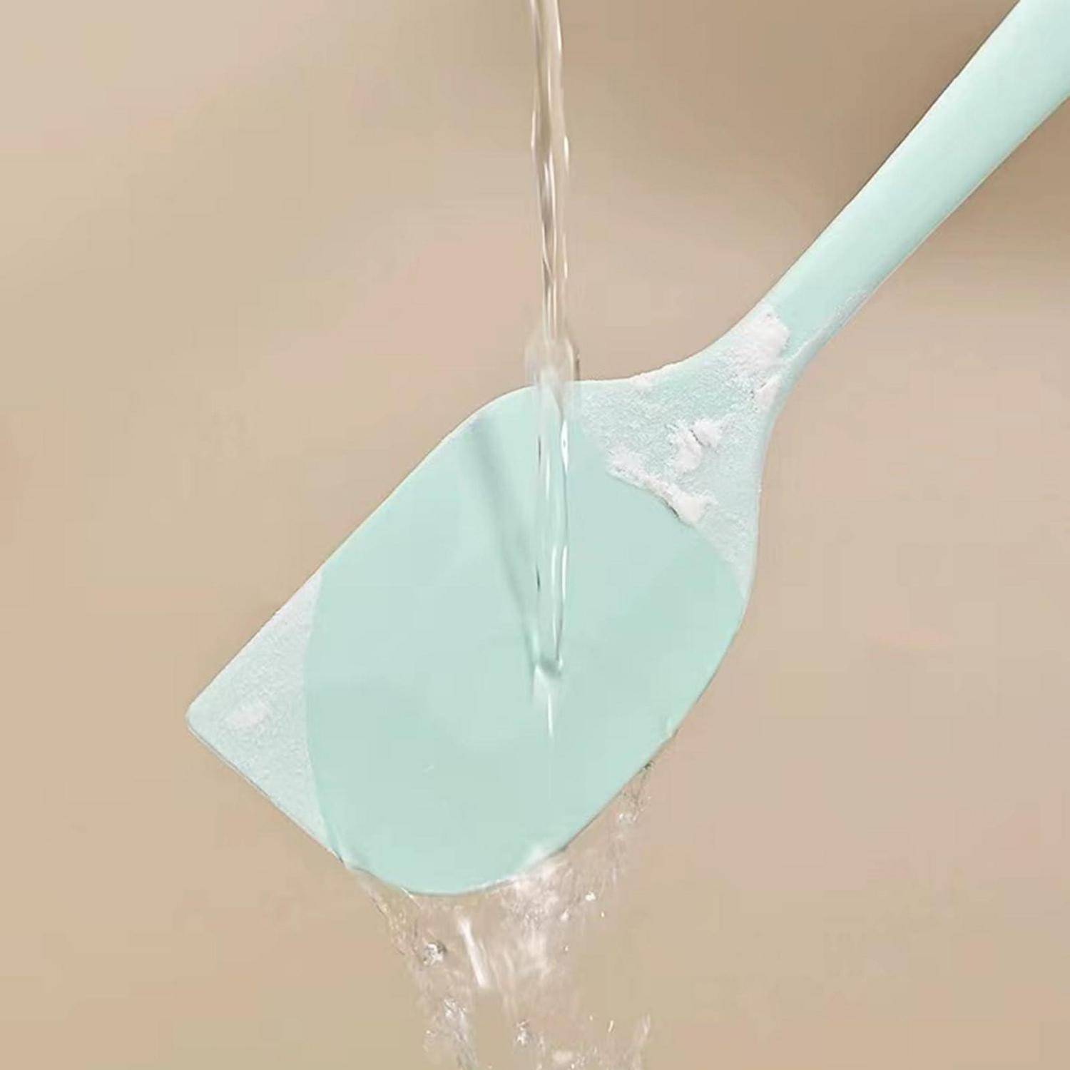 Wasser läuft über einen mintgrünen Silikonspatel und spült weiße Rückstände vor einem beigen Hintergrund ab.