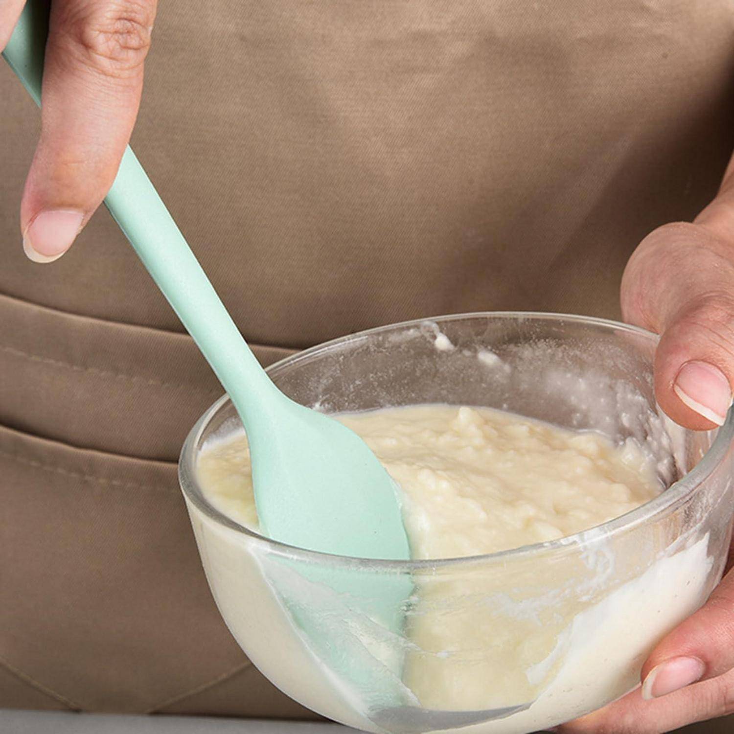 Eine Person rührt einen dicken Teig oder eine dickflüssige Masse in einer Glasschüssel mit einem mintgrünen Spatel um.