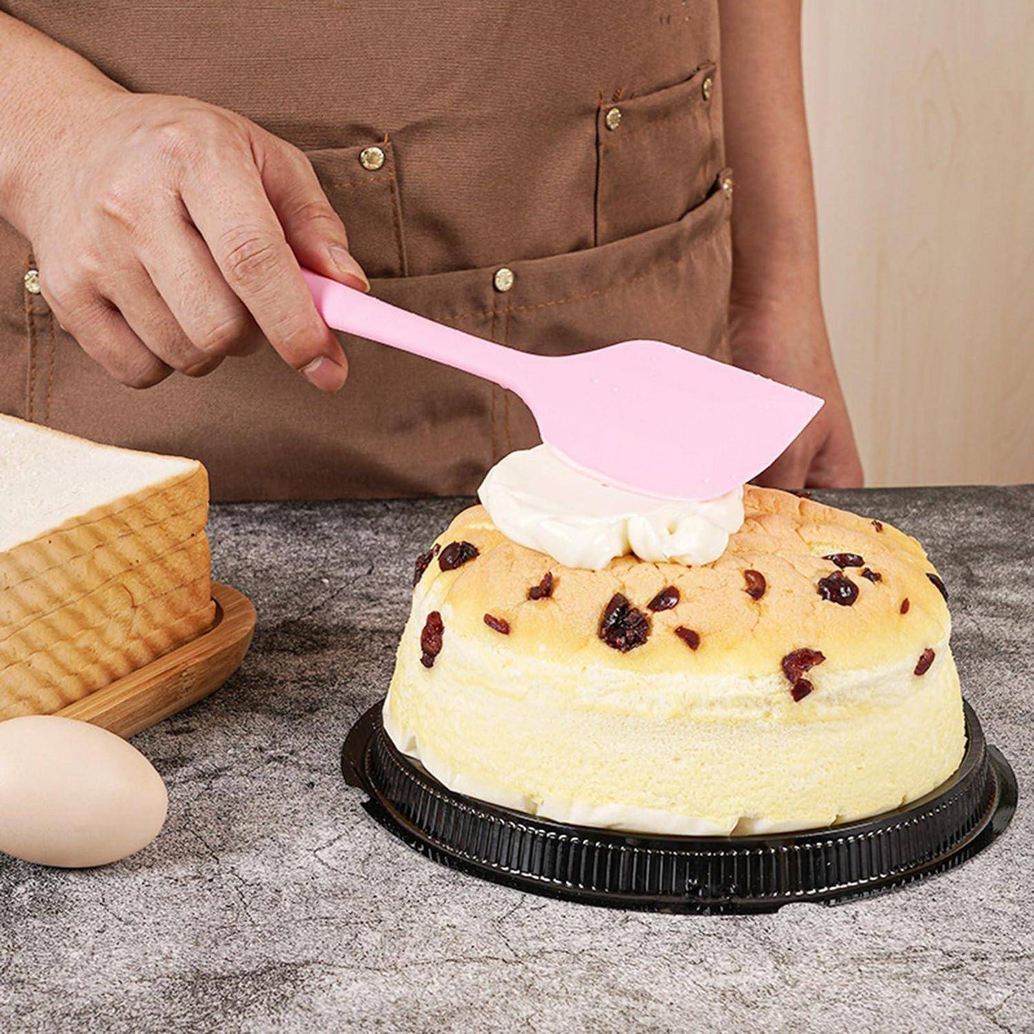 Eine Person verteilt Creme auf einem obstkuchendeckeltem Kuchen mit einem rosa Spachtel auf einer Arbeitsplatte, mit Eiern und einer Kastenform in der Nähe.