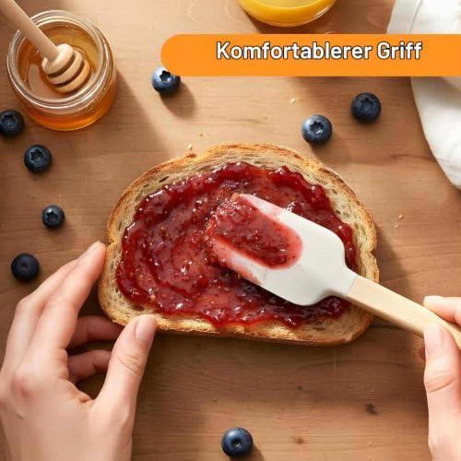 Hände, die Marmelade auf eine Brotscheibe mit einem Spachtel mit weißem Griff verteilen, mit Blaubeeren und einem Honigglas auf einem Holztisch; der Text lautet „Komfortablerer Griff