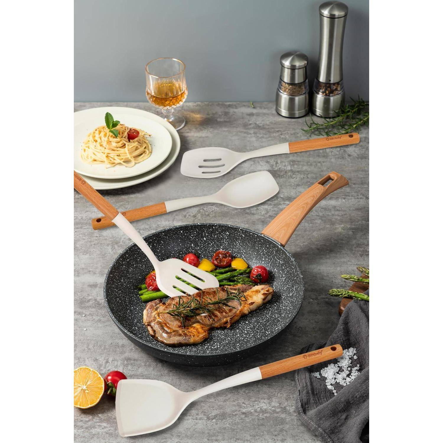 Gebratenes Steak in einer gesprenkelten Bratpfanne mit Gemüse, umgeben von Servierlöffeln und einem angerichteten Nudelgericht auf einer Küchenarbeitsplatte.