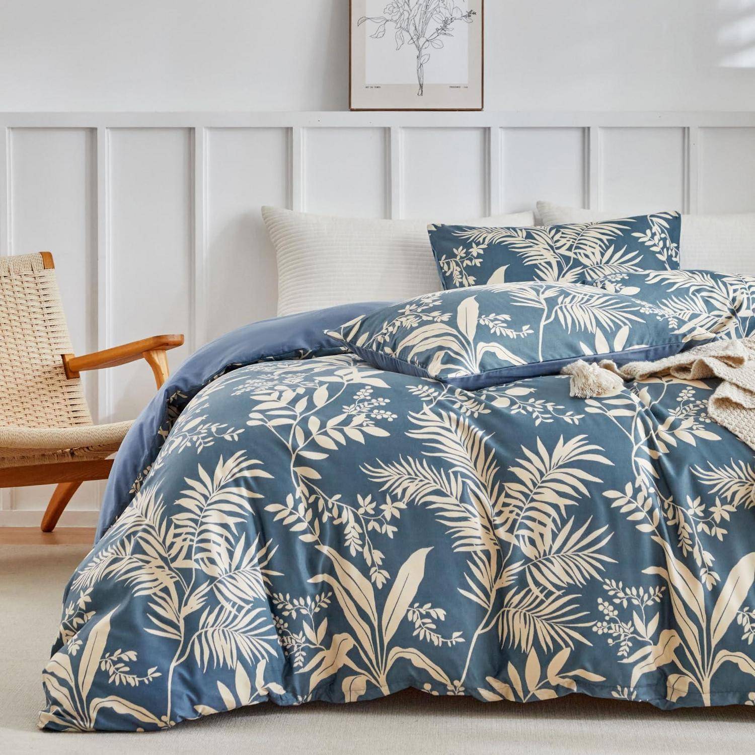 Blaue Blumen-Bettwäsche und passende Kissenbezüge auf einem ordentlich gemachten Bett in einem hellen, minimalistischen Schlafzimmer mit einem Holzstuhl und gerahmter Kunst.