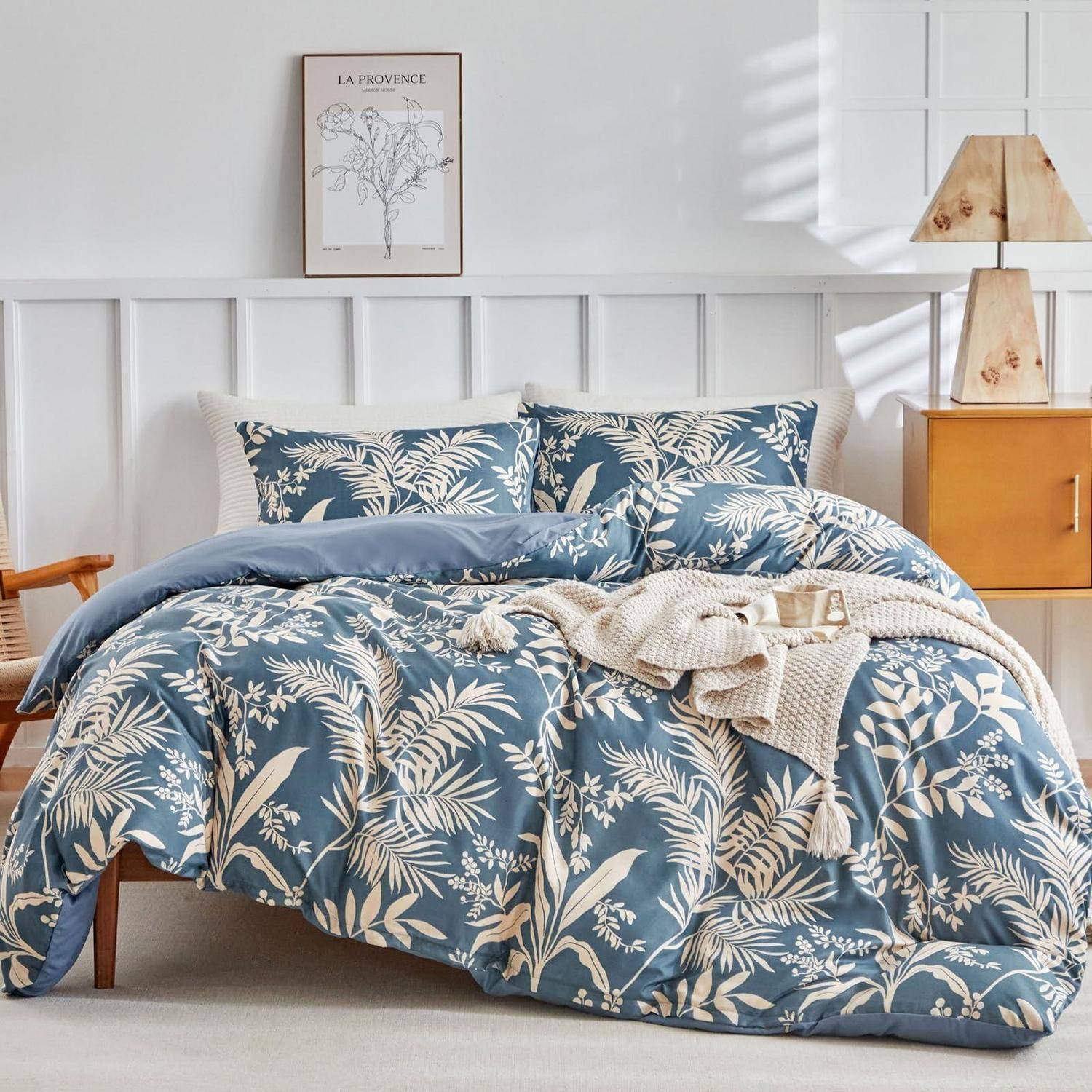 Bett mit blauem Blumenmuster mit passenden Kissen und Decke in einem hellen Schlafzimmer, mit Nachttisch, Lampe und Wandkunst.