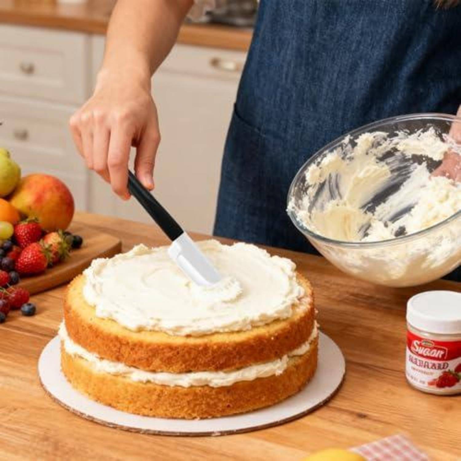 Eine Person streicht Frosting auf einen geschichteten Biskuitkuchen auf einer Küchentheke, mit einer Schüssel Zuckerguss und frischem Obst in der Nähe.