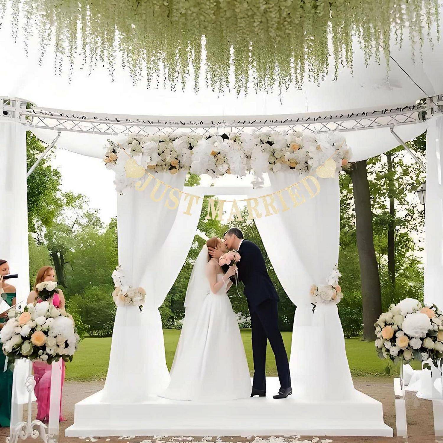 Braut und Bräutigam küssen sich auf einer dekorierten weißen Hochzeitsbühne mit Blumenbogen, "JUST MARRIED""-Schild und Outdoor-Park-Umgebung.