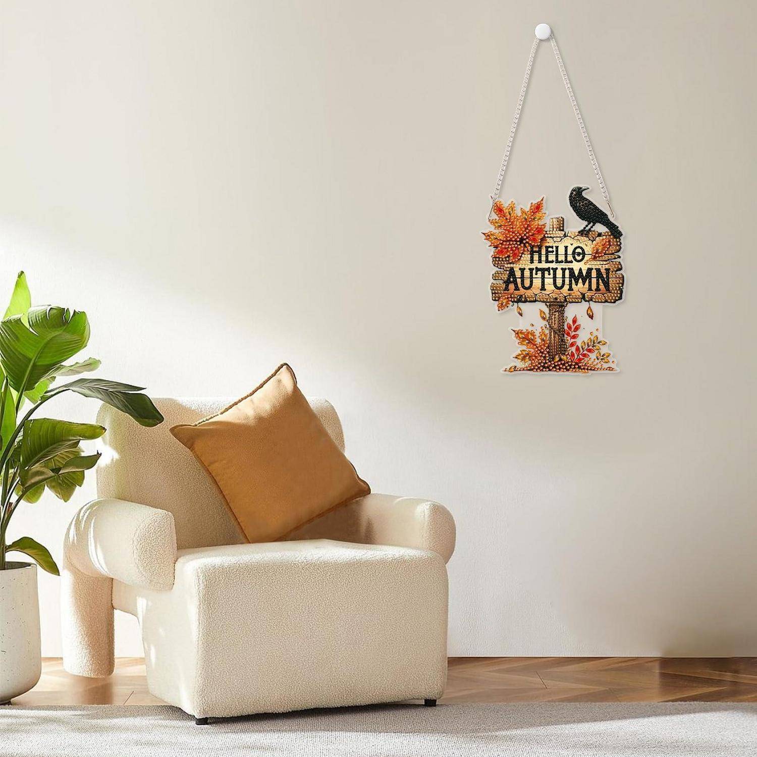 Gemütlicher beiger Sessel mit beigefarbenem Kissen neben einer Topfpflanze, unter einem hängenden Wandschild „HELLO AUTUMN" mit Herbstblättern und einem Vogel.