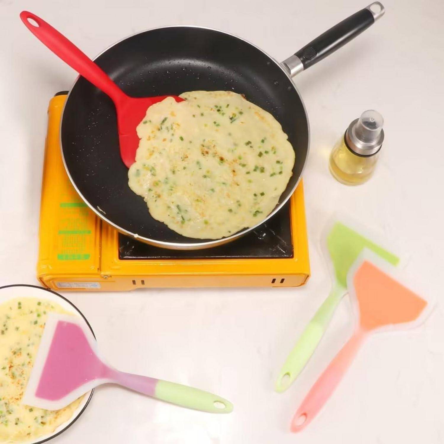 Pfannkuchen kochen in einer Pfanne auf einem tragbaren Kochfeld, mit farbigen Spachteln und einer kleinen Ölflasche in der Nähe.