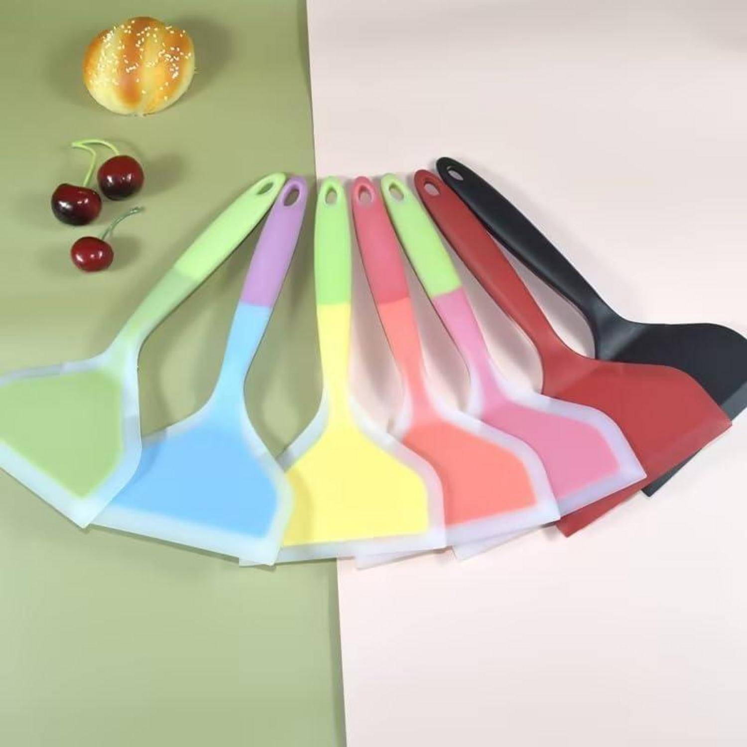 Bunte Silikonspachtel oder Wender in Fächerform auf einem geteilten grün-weißen Hintergrund angeordnet, mit einem Brötchen und Kirschen in der Nähe.