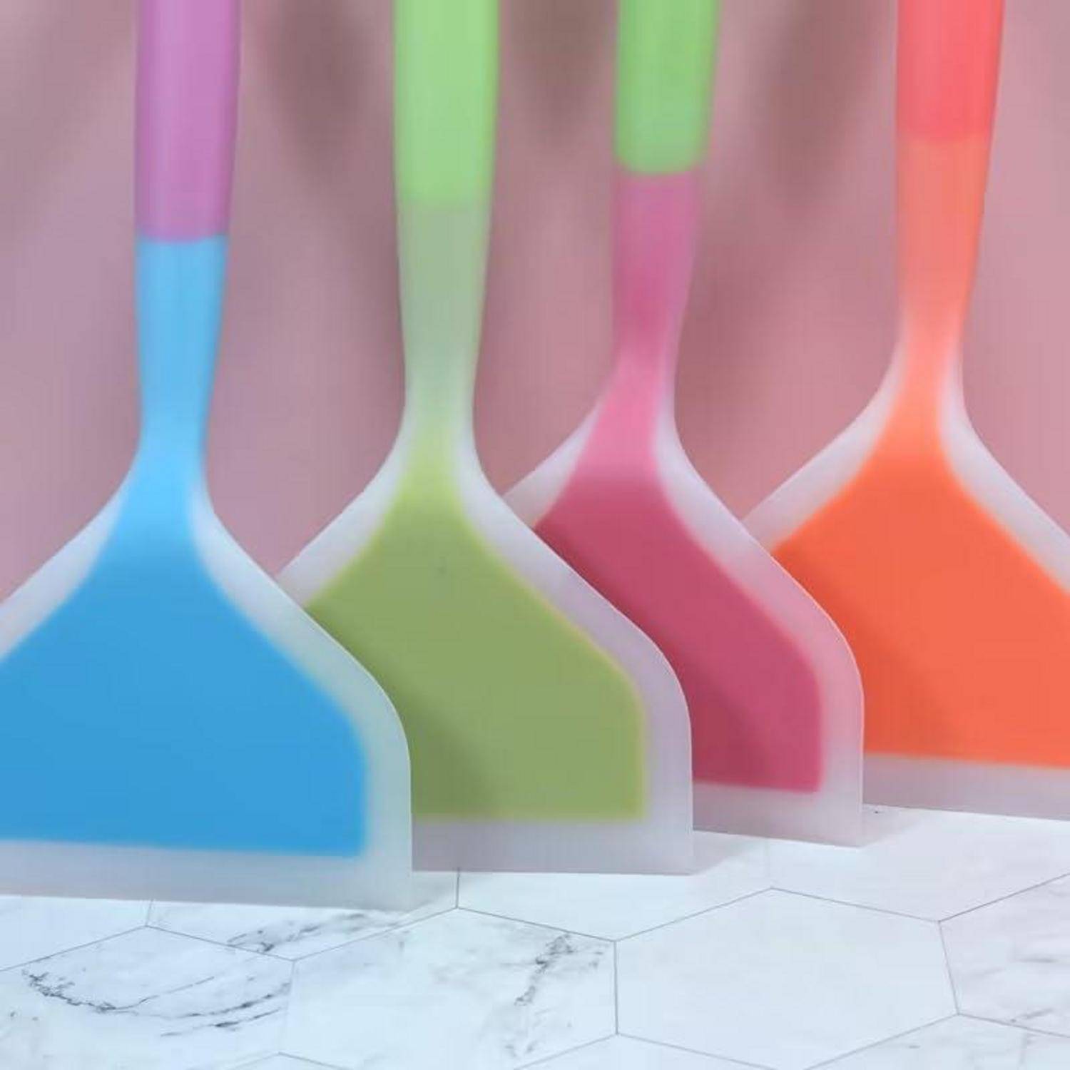 Fünf bunte Silikonspachteln, die aufrecht auf einer weißen Sechseck-Kachelfläche stehen, in Nahaufnahme gezeigt.