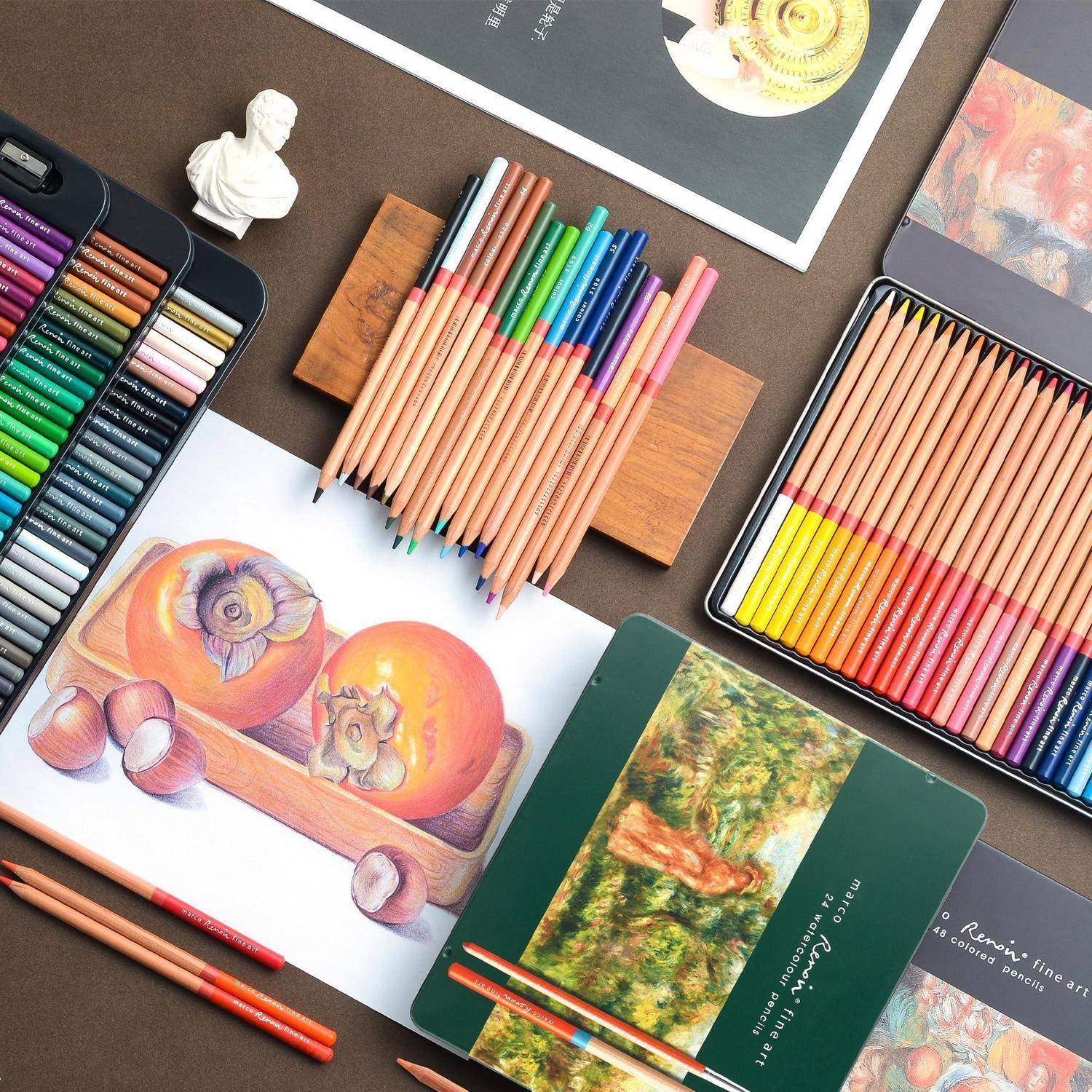 Kunstmaterialien um eine farbstiftgezeichnete Stillleben-Skizze von Obst auf einem Holztablett angeordnet, mit Farbstiften, Pastellsätzen und Skizzenbüchern auf einem Tisch.