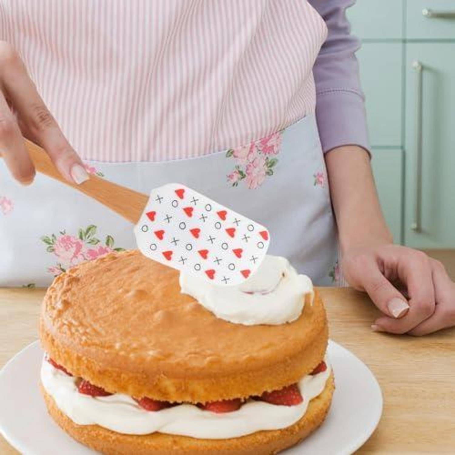 Eine Person verteilt weißes Frosting auf einem geschichteten Biskuitkuchen mit Erdbeeren und dekoriert ihn in einer Küche.