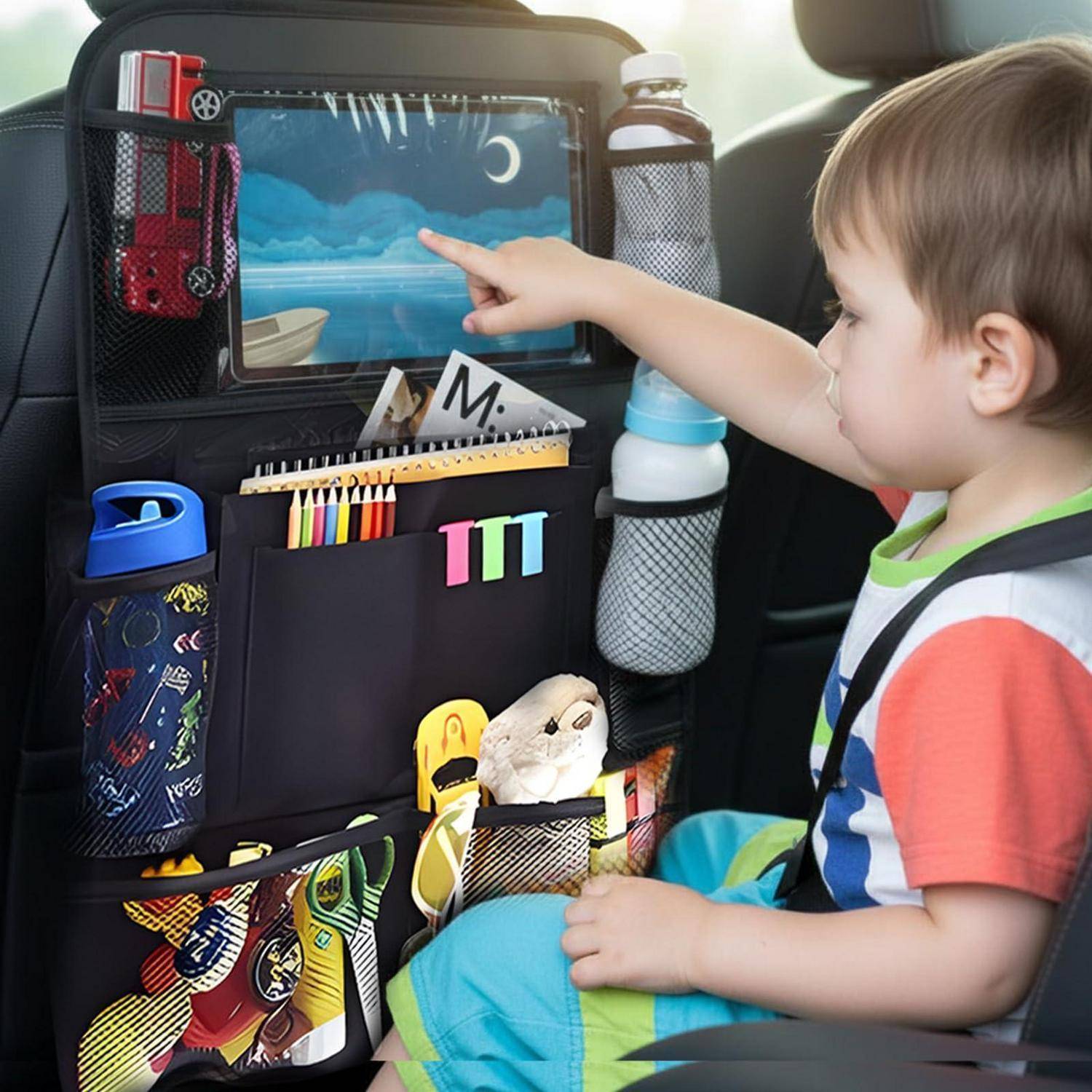 Ein kleines Kind in einem Autositz zeigt auf ein Tablet, das an der Rückenlehne des Sitzes befestigt ist, mit Reiseorganizern, Buntstiften und Wasserflaschen in der Nähe.