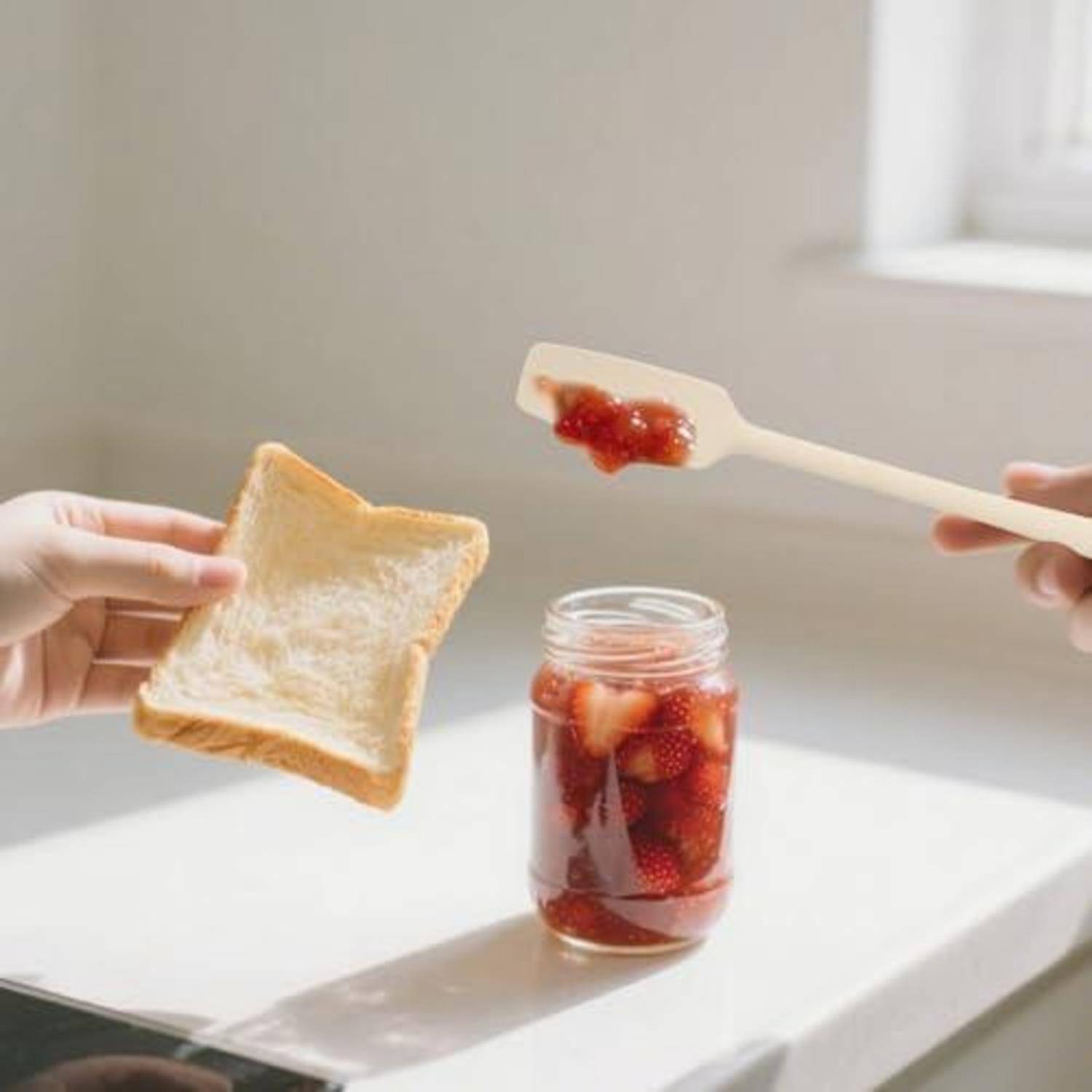 Hände bereiten ein Marmeladenbrot vor: eine hält eine Scheibe Toast, während die andere Erdbeermarmelade aus einem Glas streicht.