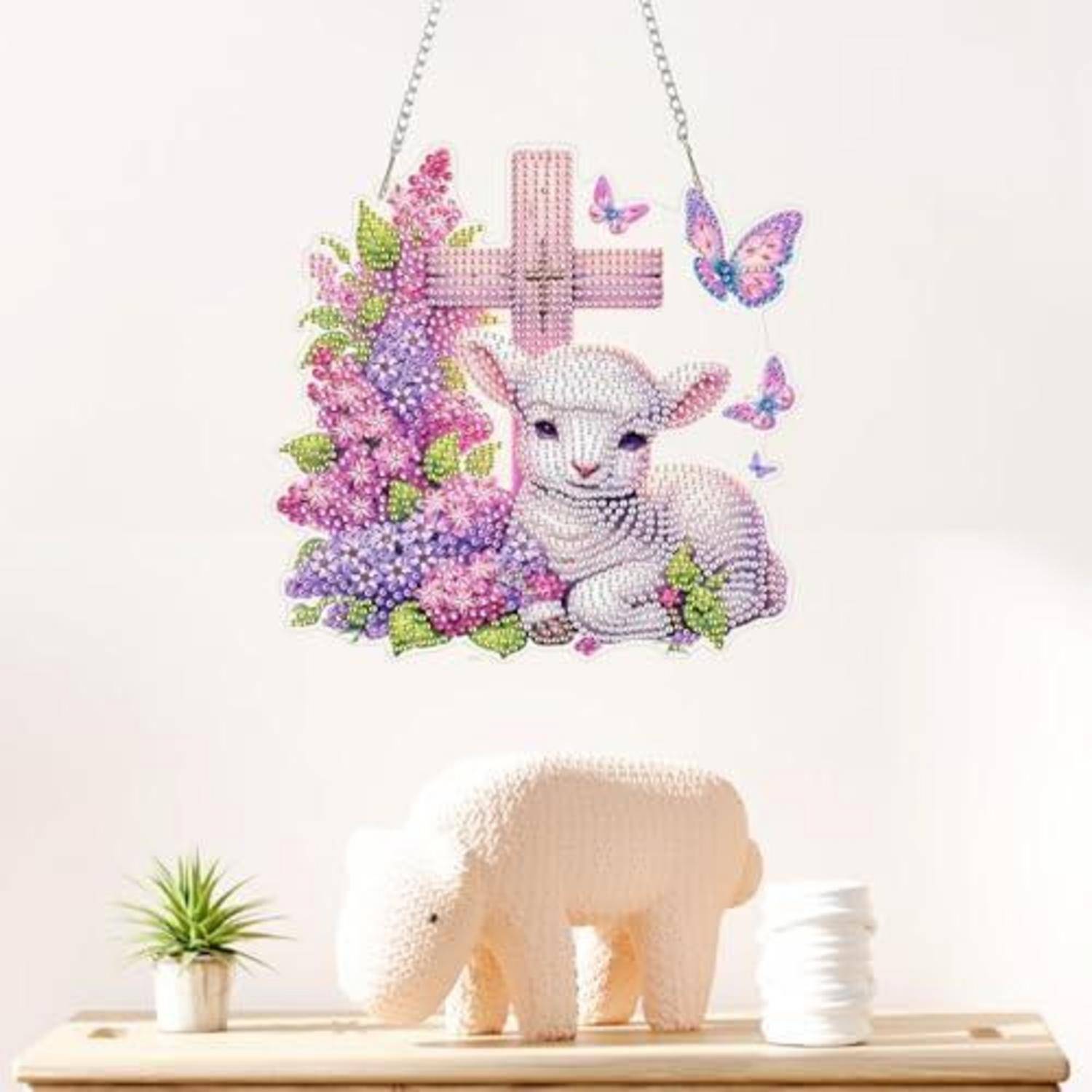 Eine Wanddekoration zum Aufhängen zeigt ein Lamm neben violetten Blumen und Schmetterlingen, mit einem rosa Kreuz darüber. Auf einem Regal darunter sitzt eine weiße bärenförmige Skulptur neben einer kleinen getopften Pflanze und einem Stapel weißer Gegenstände.