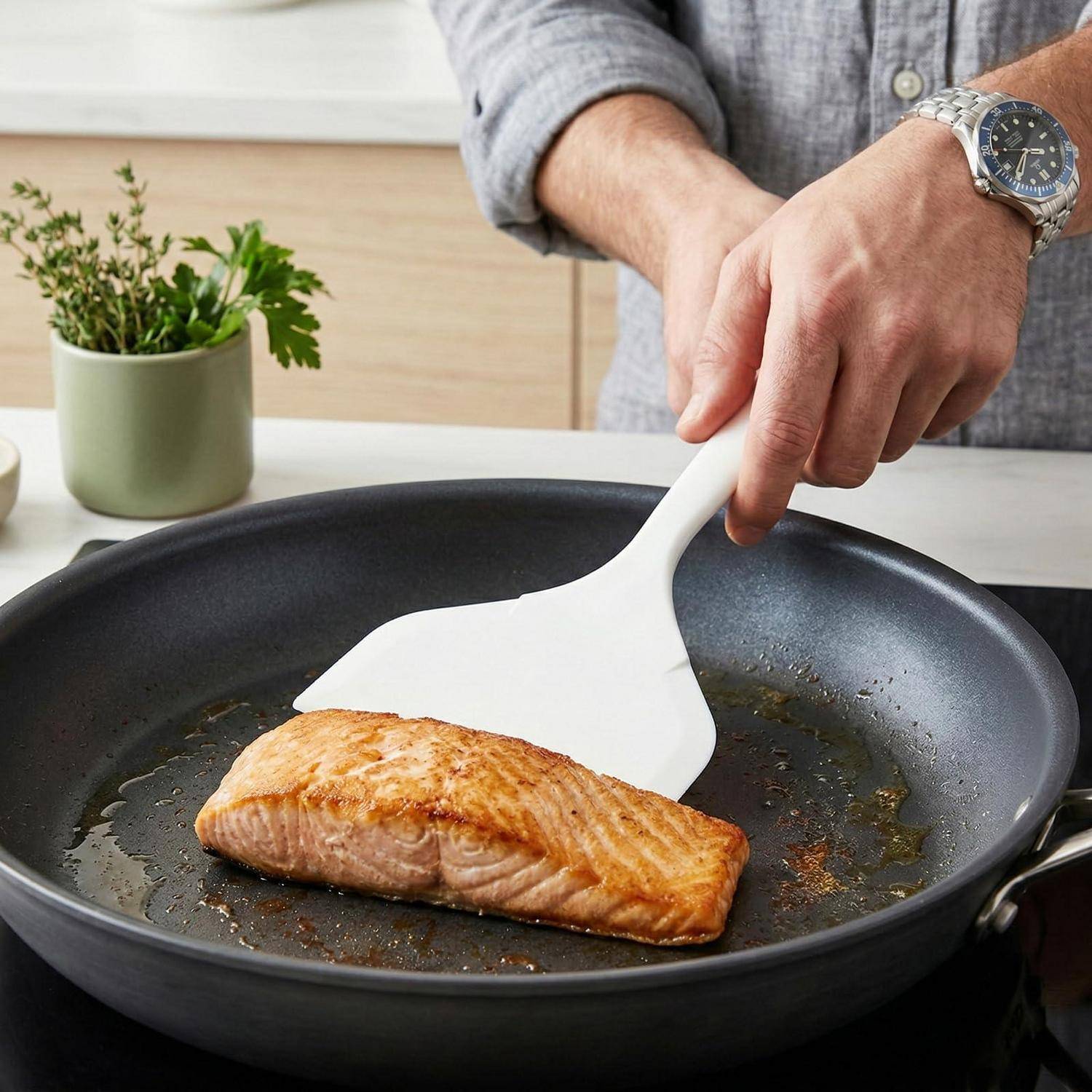Eine Person verwendet einen weißen Spatel, um ein angebratenes Lachsfilet in einer schwarzen Bratpfanne auf einer Küchenarbeitsplatte zu wenden.