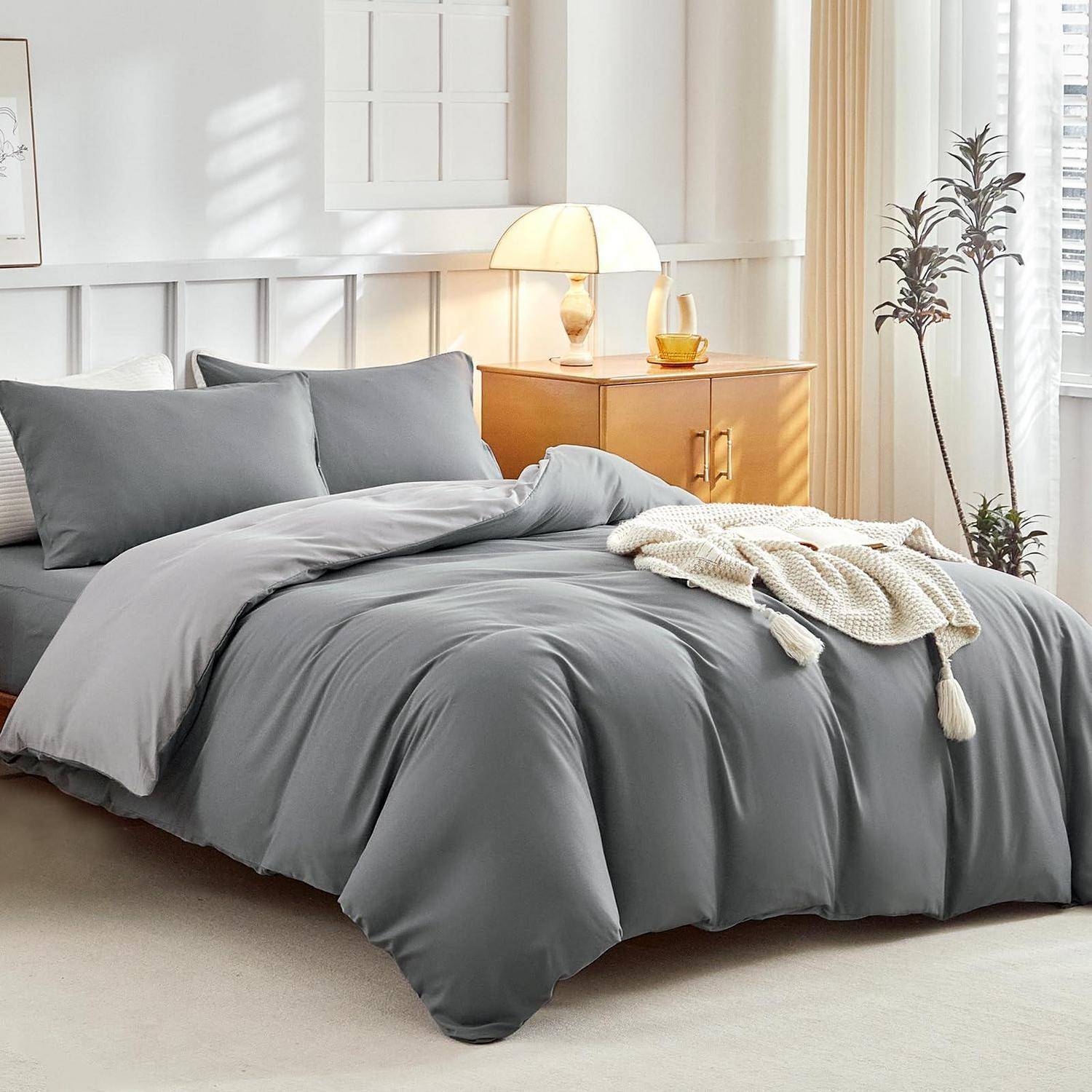 Graue Bettwäsche auf einem ordentlich gemachten Bett in einem hellen Schlafzimmer, mit Kissen, einer Wurfdecke und einer Nachttischlampe auf einem Holznachttisch.