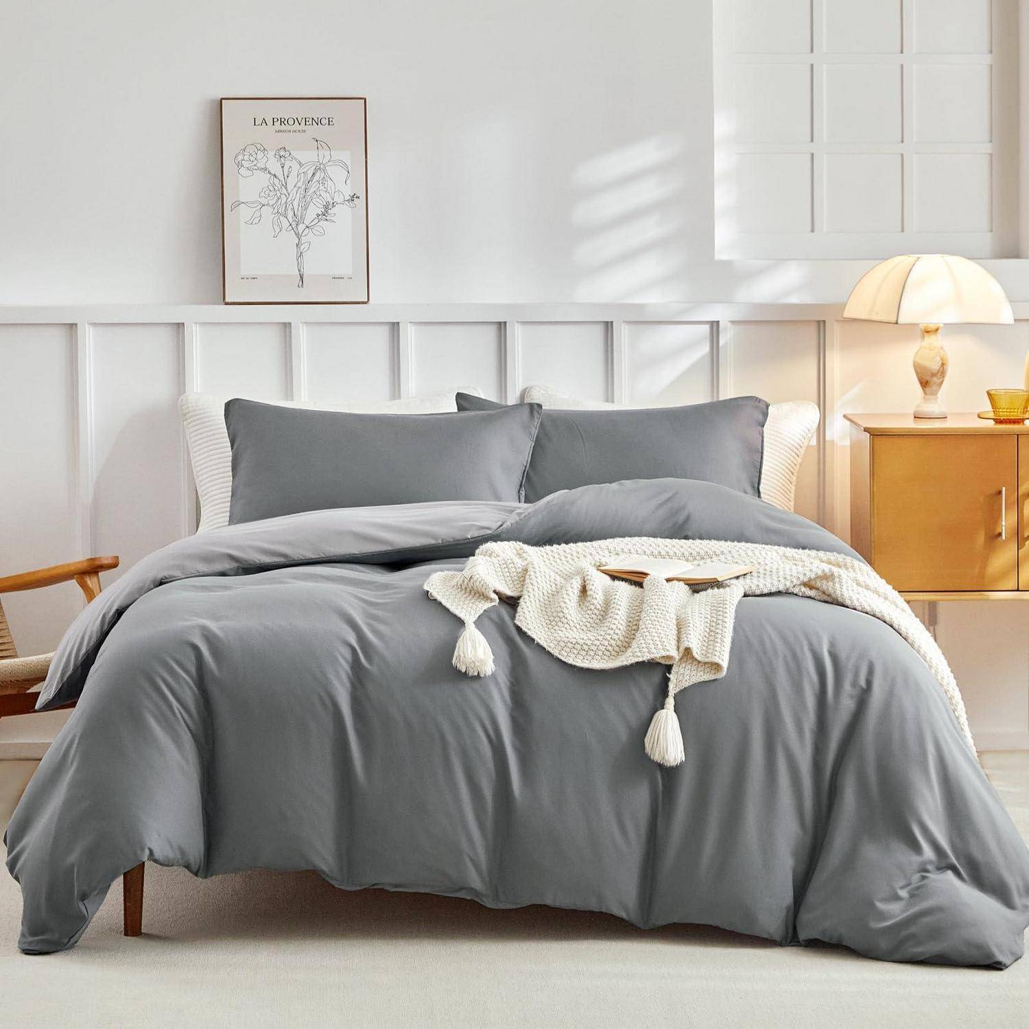 Ein ordentlich gemachtes Bett mit grauer Bettwäsche in einem hellen, minimalistischen Schlafzimmer mit einer Lampe auf einem Nachttisch und gerahmter Wandkunst über dem Kopfende des Bettes.