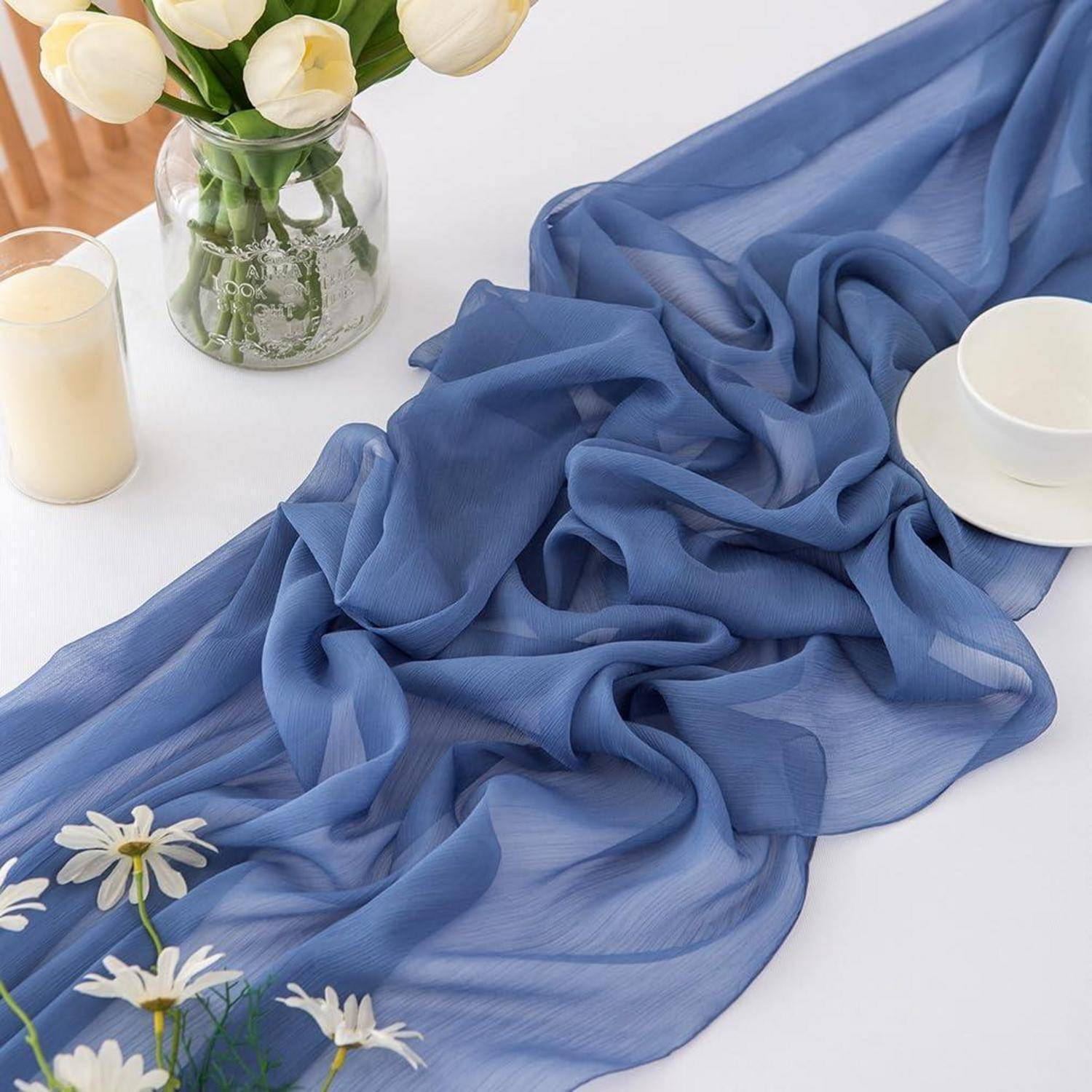 Zartes blaues Tuch, das über eine weiße Oberfläche drapiert ist, gestylt mit weißen Tulpen, einer Kerze, einer Glasvase und einer Tasse.