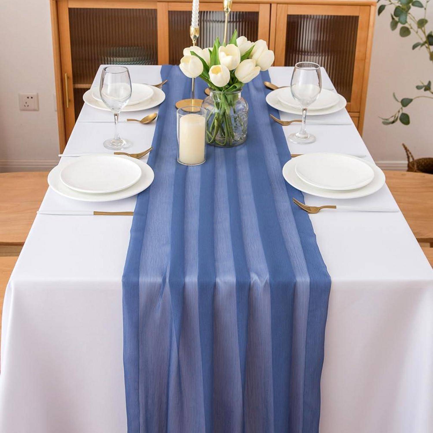 Decken Sie den Esstisch für zwei Personen mit weißen Tellern, Weingläsern, goldenem Besteck, blauem Tischläufer und einer Vase mit weißen Tulpen und Kerzen.