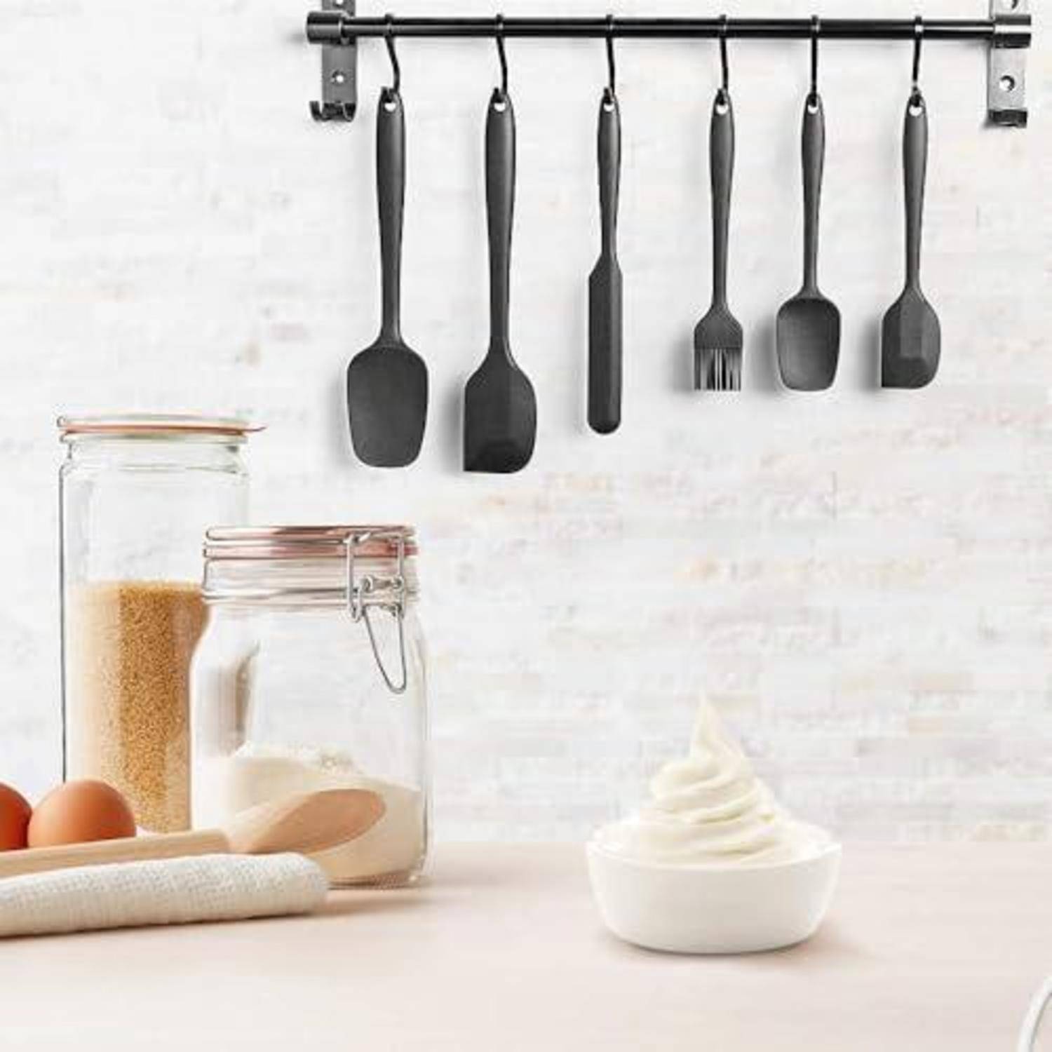 Küchenutensilien, die an einer Schiene über Gläsern mit trockenen Zutaten hängen, Eier, ein Nudelholz und eine Schüssel mit Schlagcreme auf einer Arbeitsplatte.