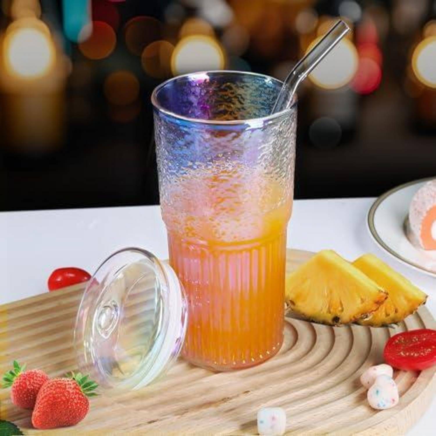 Ein großes, frostiges orangefarbenes Getränk mit einem Metallstrohhalm steht auf einem Tablett, umgeben von Erdbeeren, Ananas, Kirschtomaten und einem Deckel.