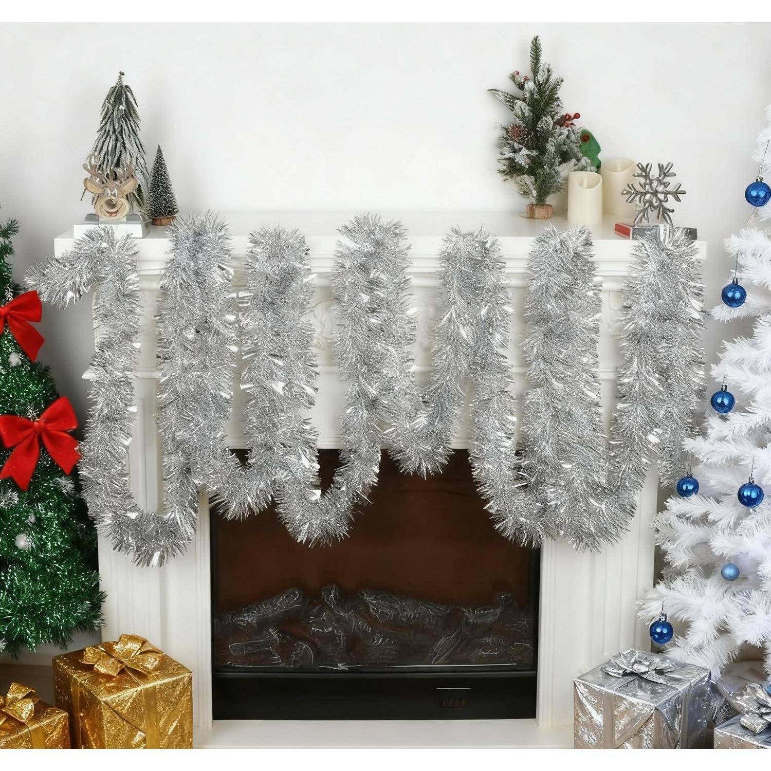 Weihnachtskamin dekoriert mit silbernen Lamettagirlanden, flankiert von einem grünen Baum mit roten Schleifen und einem weißen Baum mit blauen Kugeln und Geschenken.