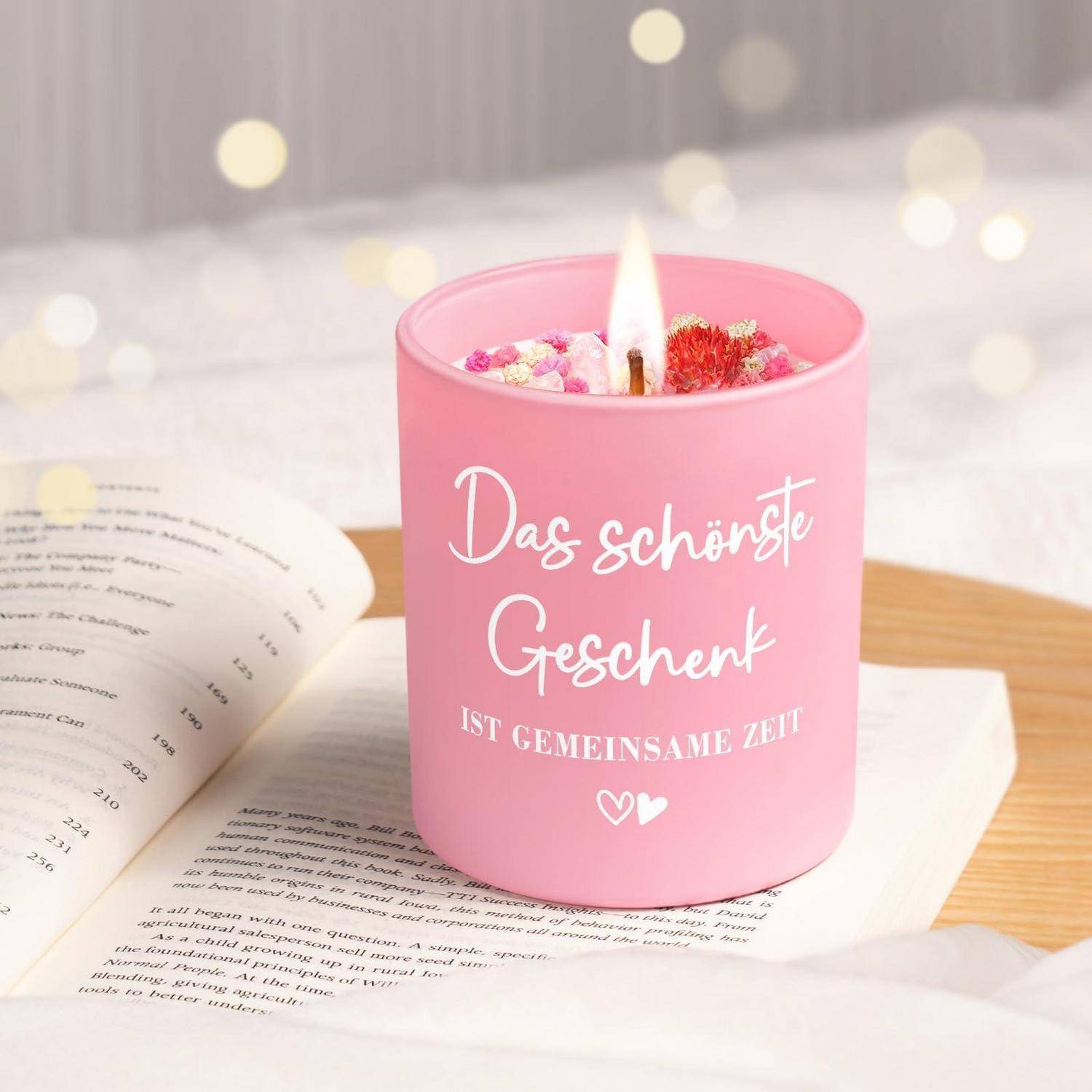 Rosa Kerze auf einem aufgeschlagenen Buch, beschriftet mit „Das schönste Geschenk ist gemeinsame Zeit", mit einer brennenden Flamme und dekorativen Blumen, was auf ein Geschenk oder eine gemütliche Leseszene hindeutet.