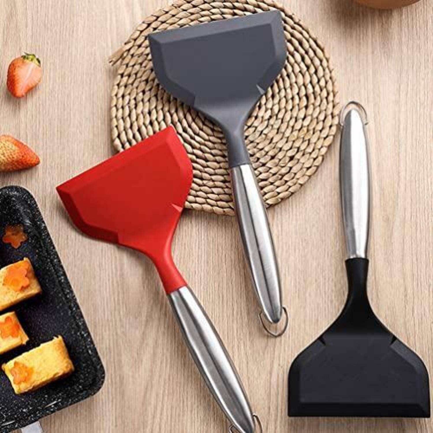 Drei Spachtel mit Metallgriffen und roten, grauen und schwarzen Köpfen auf einem Tisch neben einem geflochtenen Topfuntersetzer und kleinen Lebensmittelstücken auf einem Tablett angeordnet.