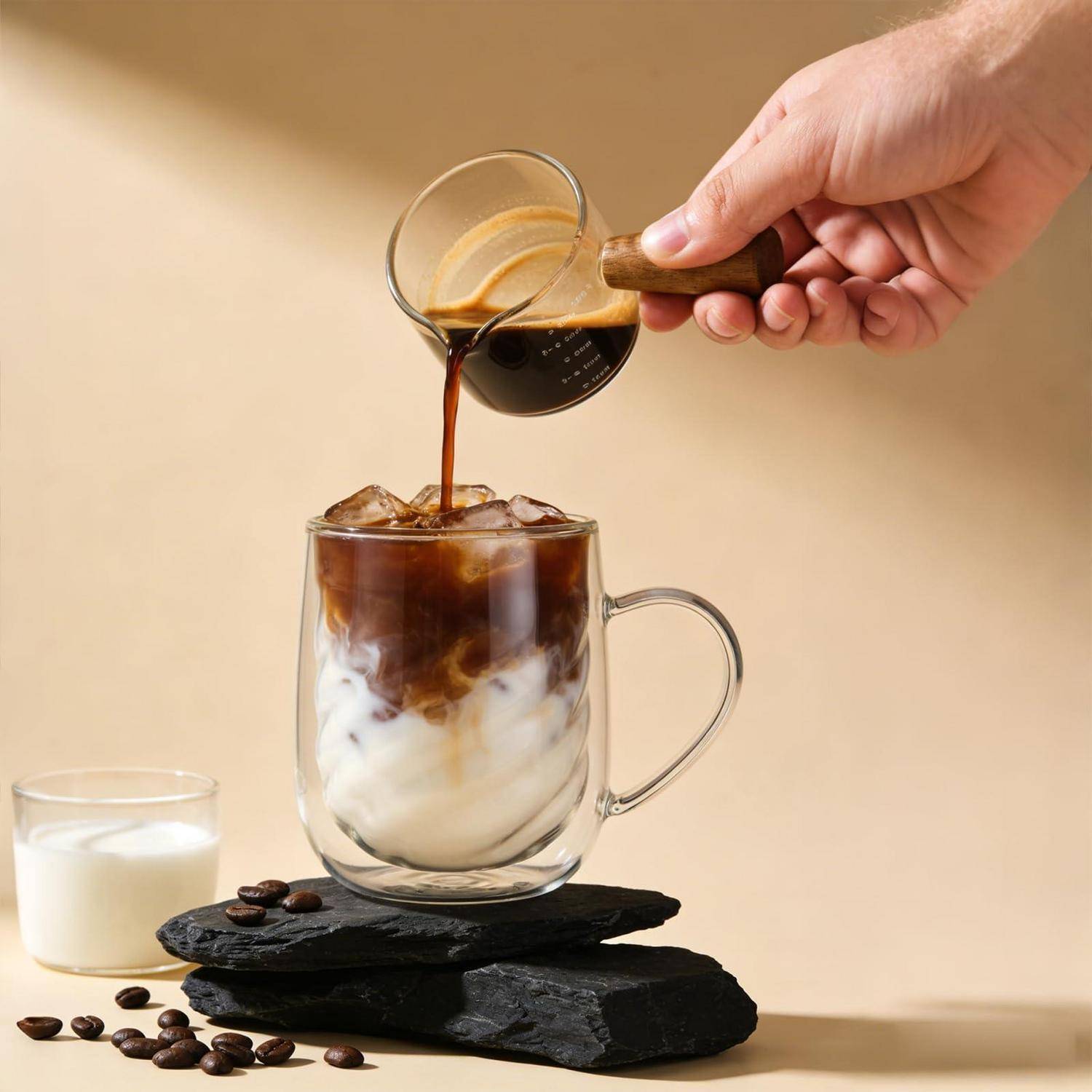Hand gießt Espresso über Eis und Milch in einer Glastasse und bereitet einen Eiskaffee auf gestapelten Schiefertafeln mit verstreuten Kaffeebohnen zu.