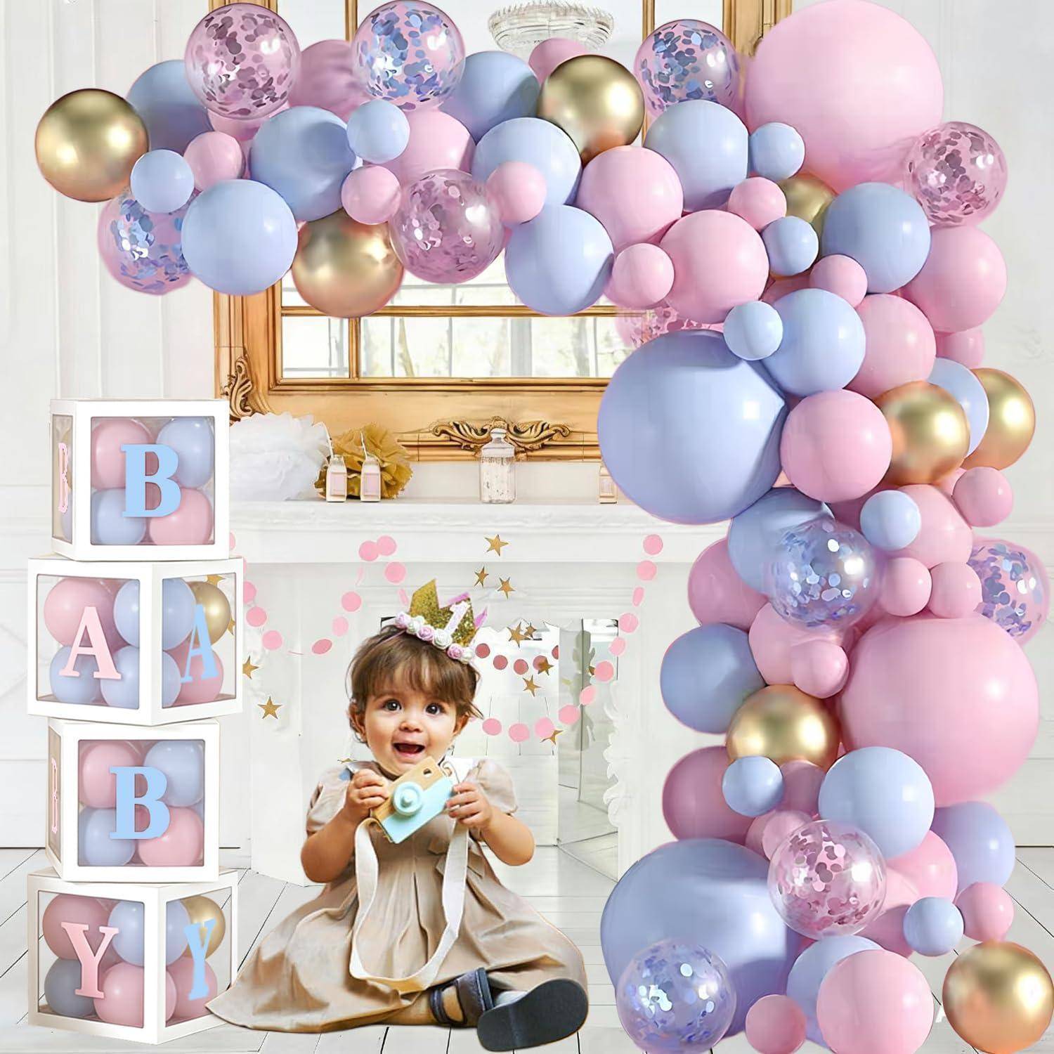 Kleines Mädchen sitzt lächelnd in einer dekorierten Erst-Geburtstags-Kulisse mit Pastellballon-Bogen, gestapelten Blöcken mit der Aufschrift „BABY" und einem Kuchendeko.